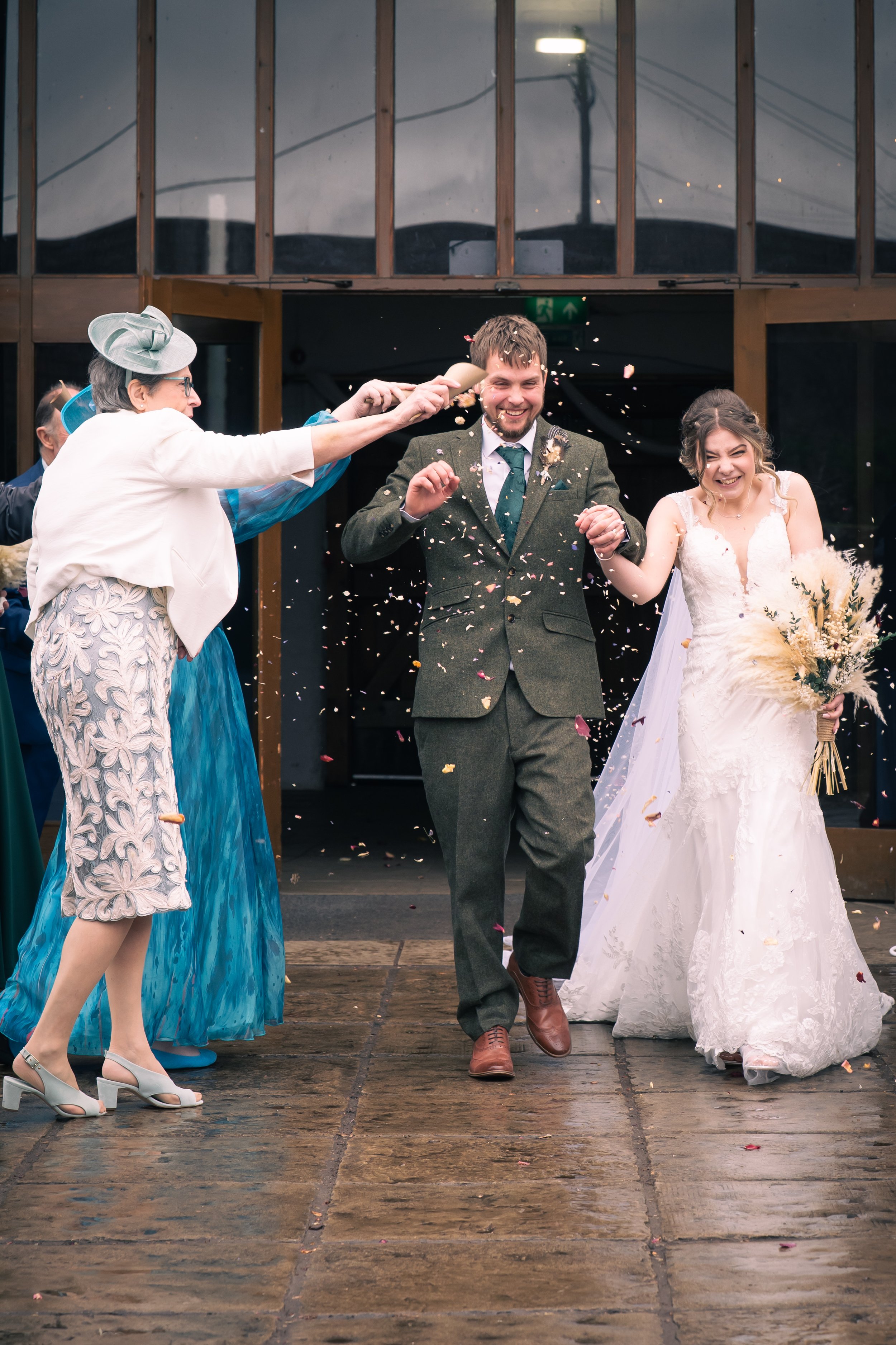 A newlywed couple walking out of a building, holding hands, as an older woman throws confetti at them. The groom is wearing a green suit with a tie, and the bride is in a white wedding dress holding a bouquet. Everyone seems joyful, celebrating their