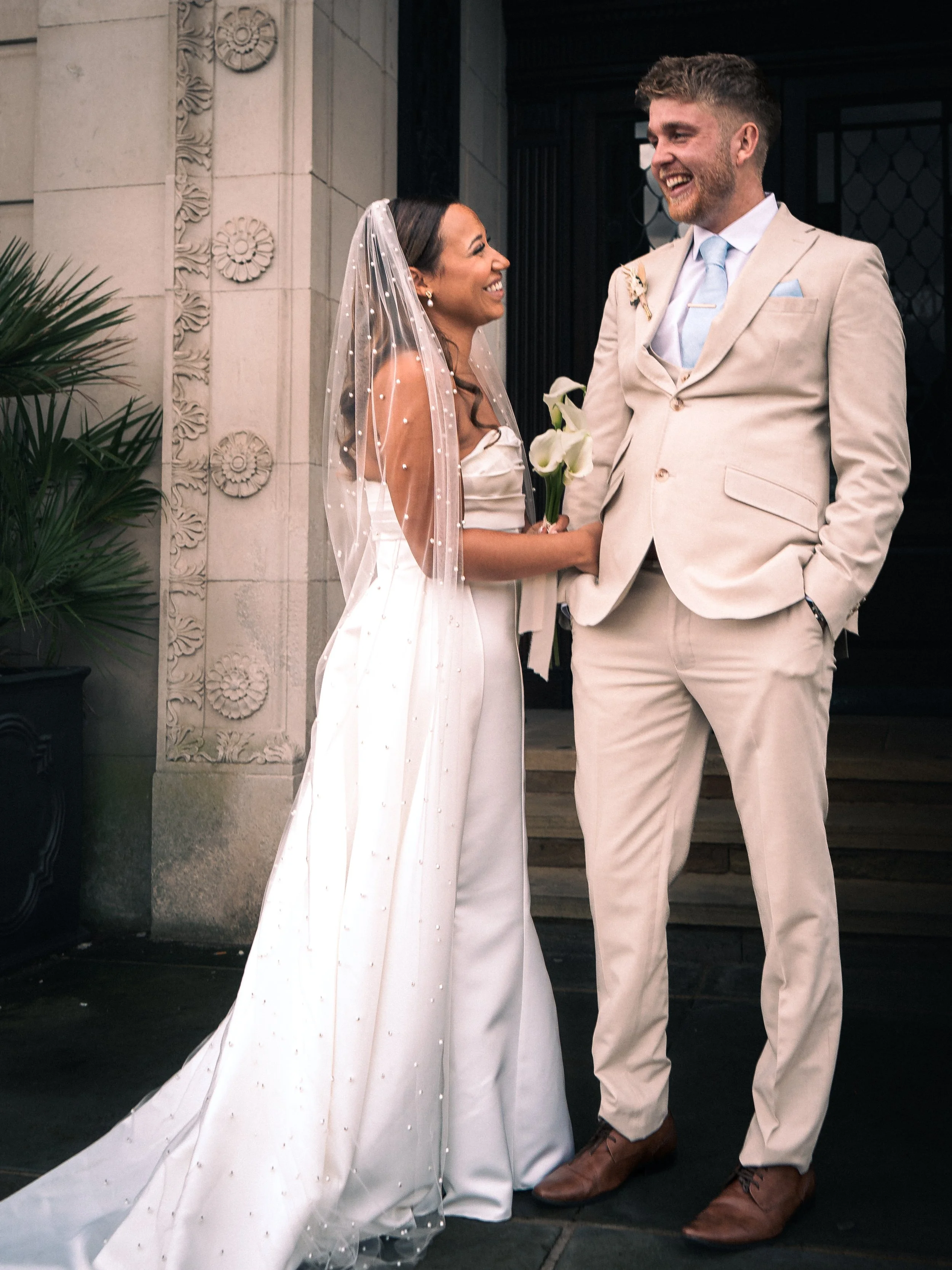 A bride and groom standing outside a building, smiling at each other, holding hands. The bride is wearing a white wedding gown and veil, holding a white calla lily. The groom is wearing a cream-colored suit with a light blue tie and pocket square.