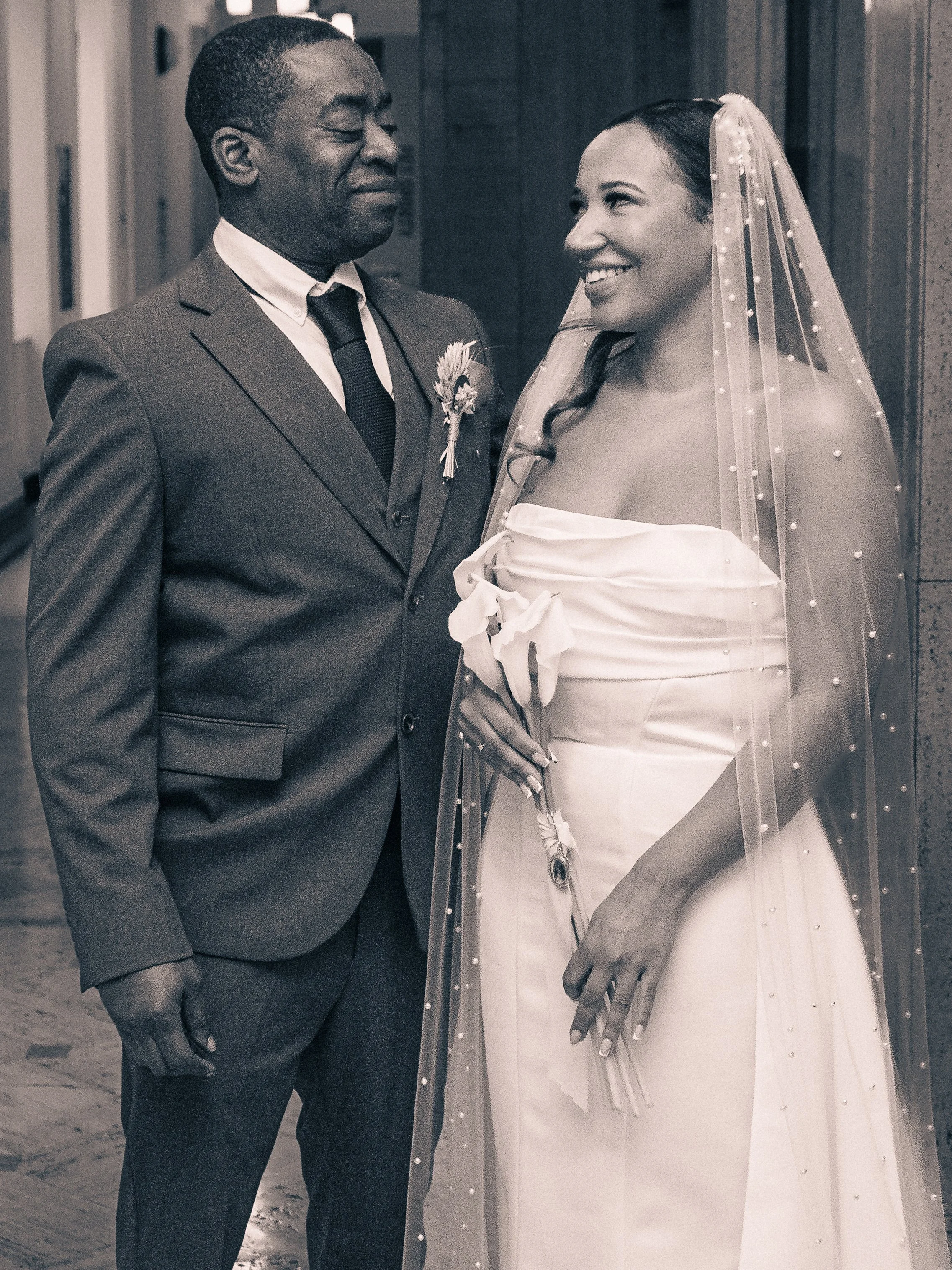 A joyful bride and an elegant groom share a tender moment on their wedding day, standing indoors with the bride wearing a veil and a strapless white wedding dress, and the groom in a suit with a boutonniere.