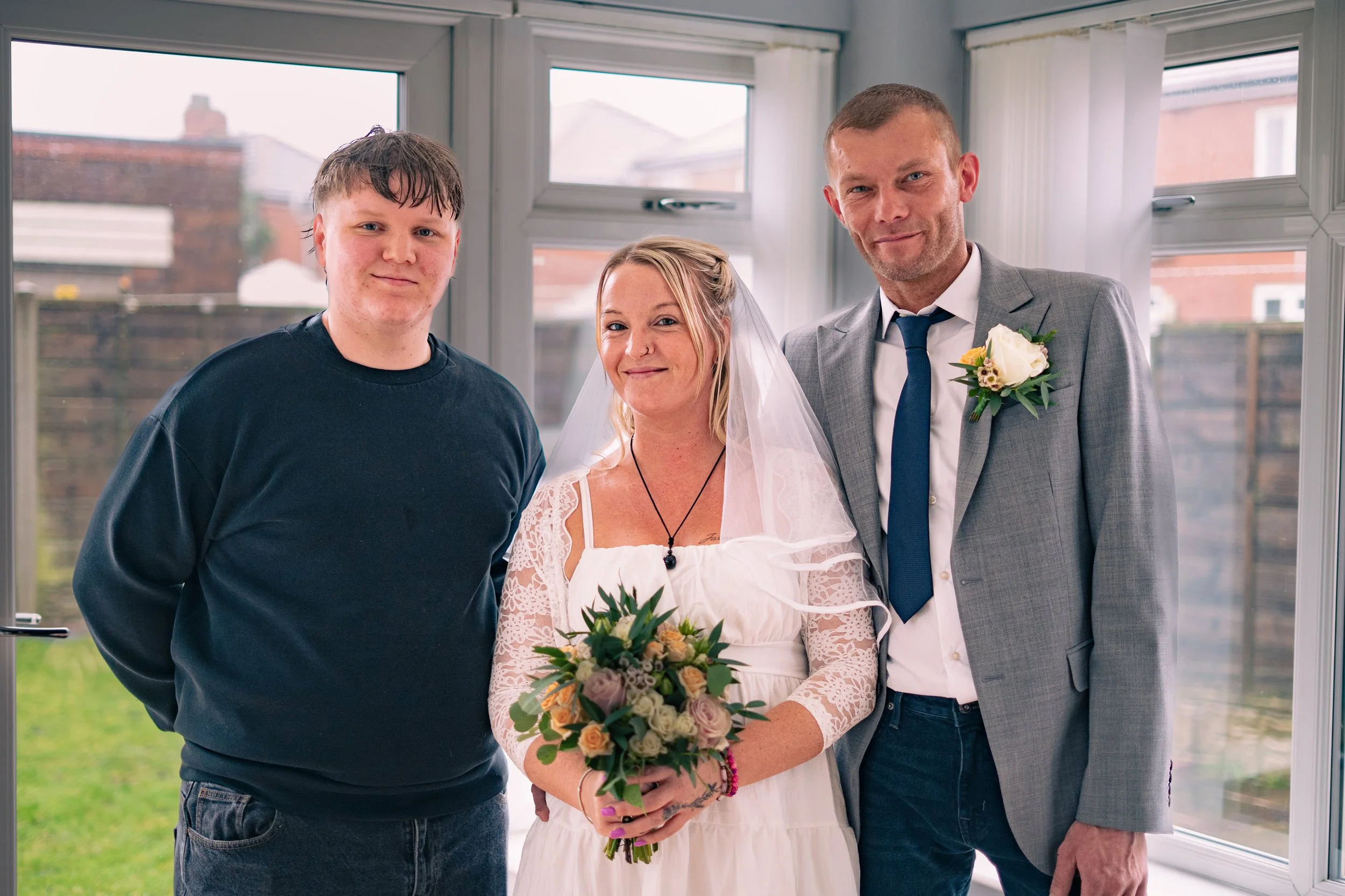 Three people at a wedding: a young man on the left, a woman in the center holding a bouquet, and a man in a suit on the right, standing indoors near windows.