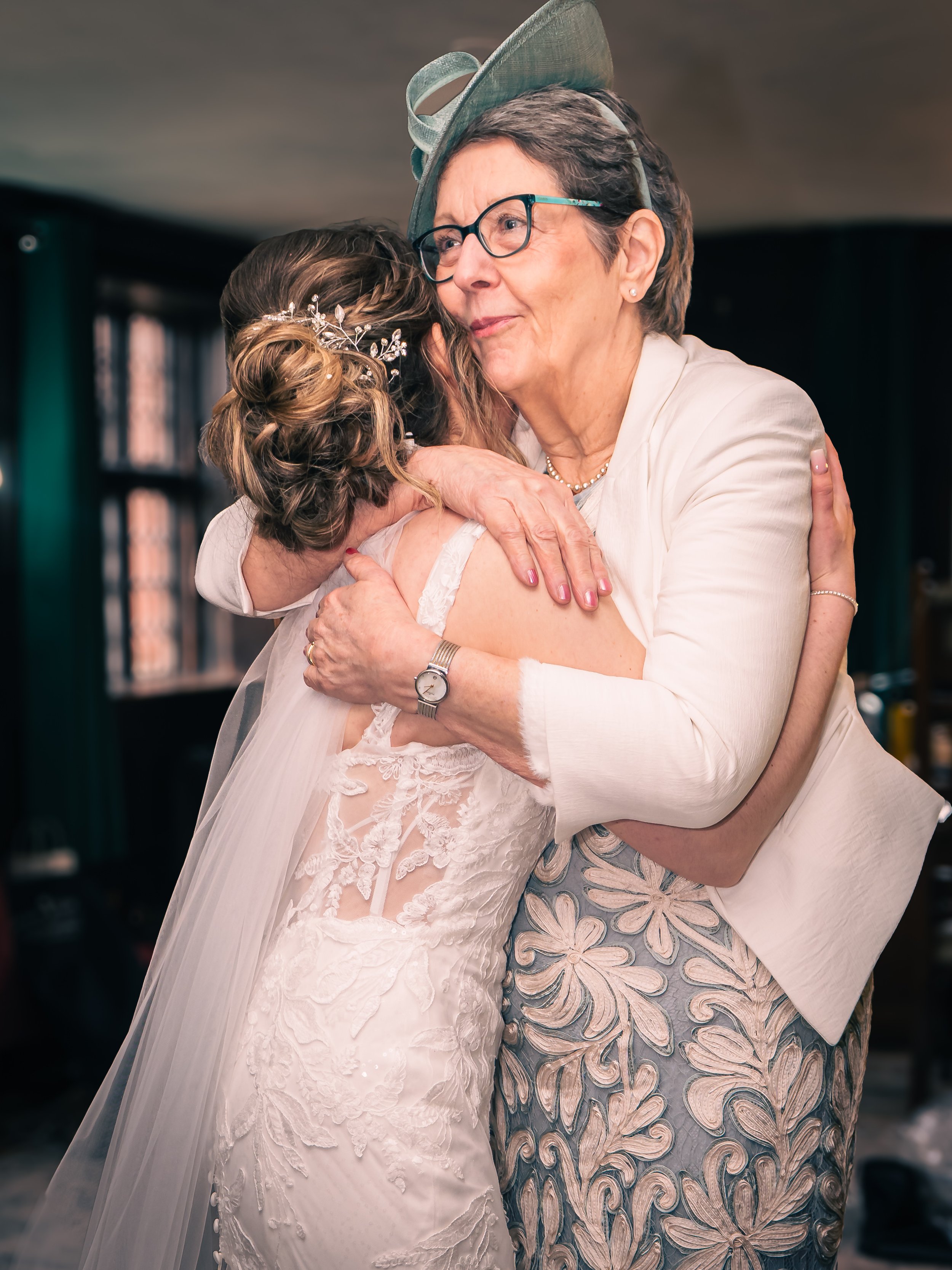 A young bride and an older woman, likely her mother or grandmother, are hugging warmly at a wedding. The bride is wearing a lace wedding dress and has her hair styled with a decorative headpiece. The older woman is wearing glasses, a light-colored bl