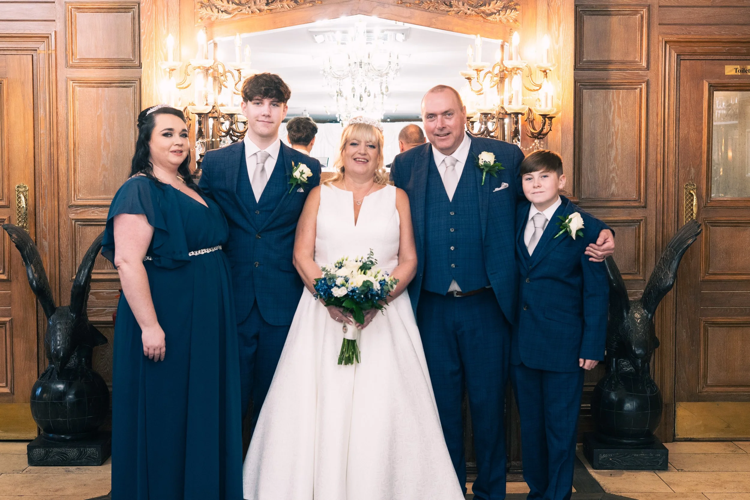 Group of wedding family members standing together indoors. The bride in a white dress holding a bouquet, surrounded by four other people in blue suits and dresses, with wood-paneled walls and decorative lighting in the background.