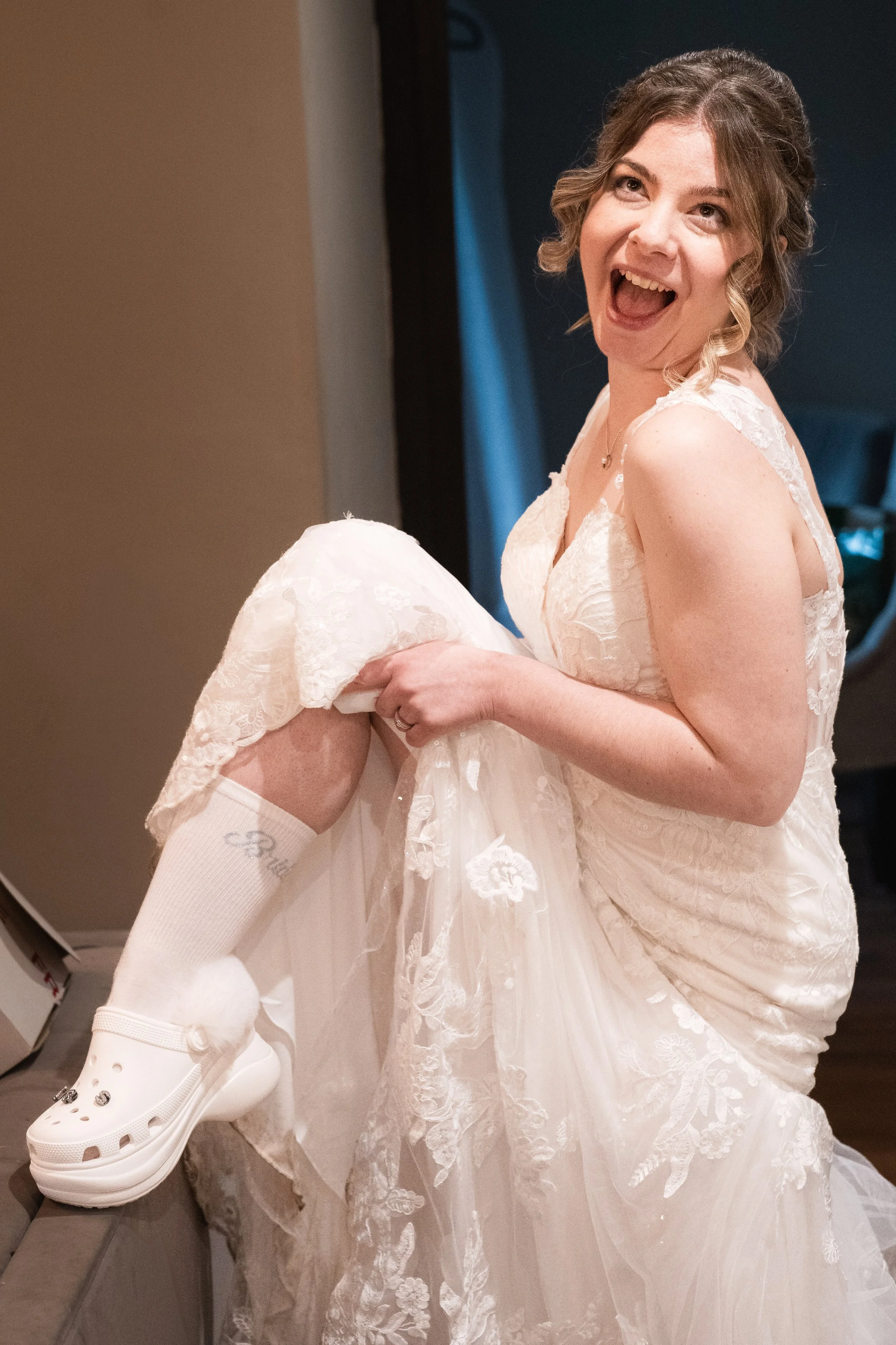 A woman in a wedding dress smiling and lifting her dress to reveal a white knee-high sock with a name and a white Croc shoe with a pom-pom, inside a room.
