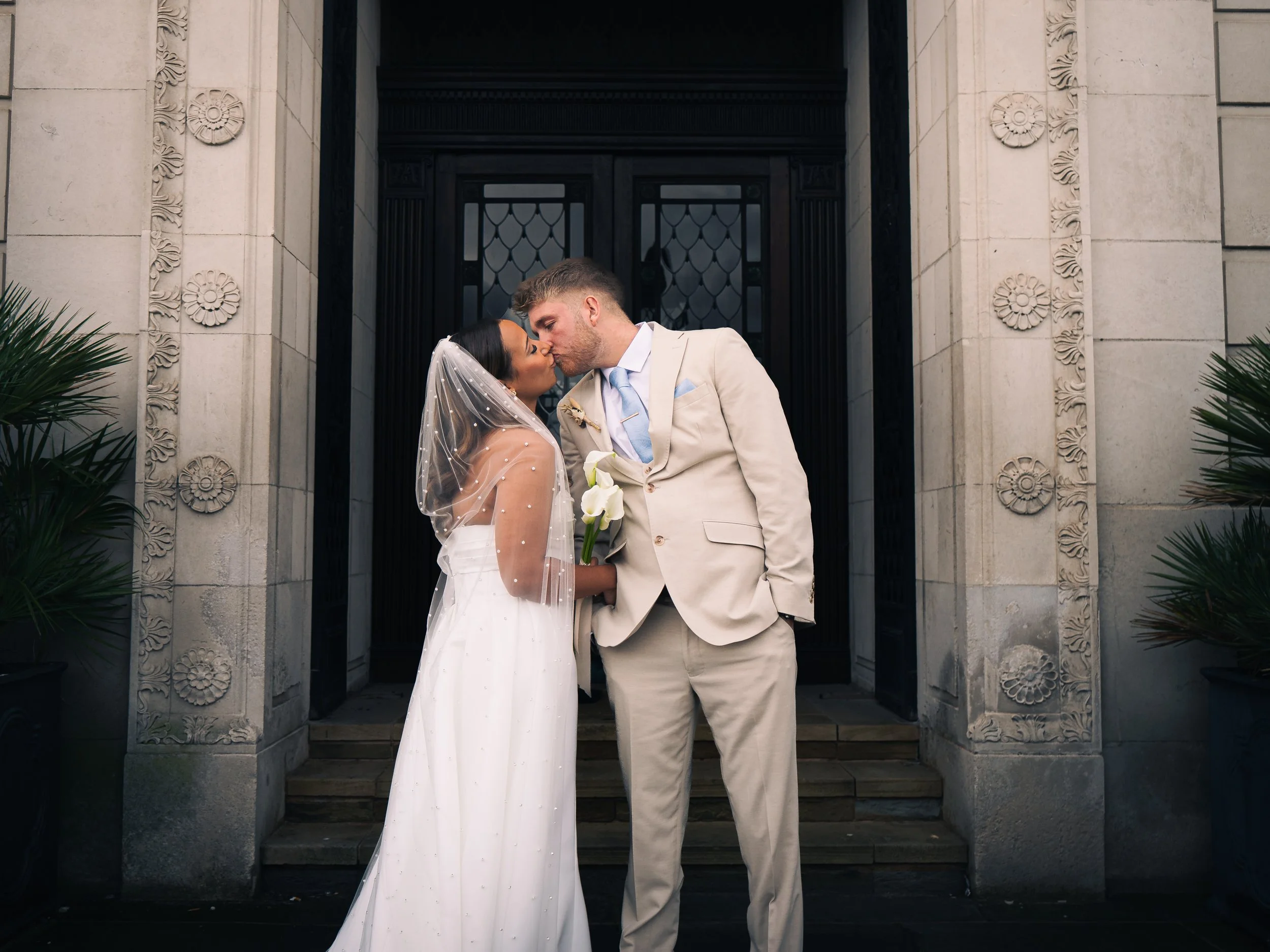 A newlywed couple sharing a kiss outside a building decorated with floral carvings, the bride wearing a white wedding dress and veil, holding a white calla lily, and the groom in a light beige suit with a light blue tie.