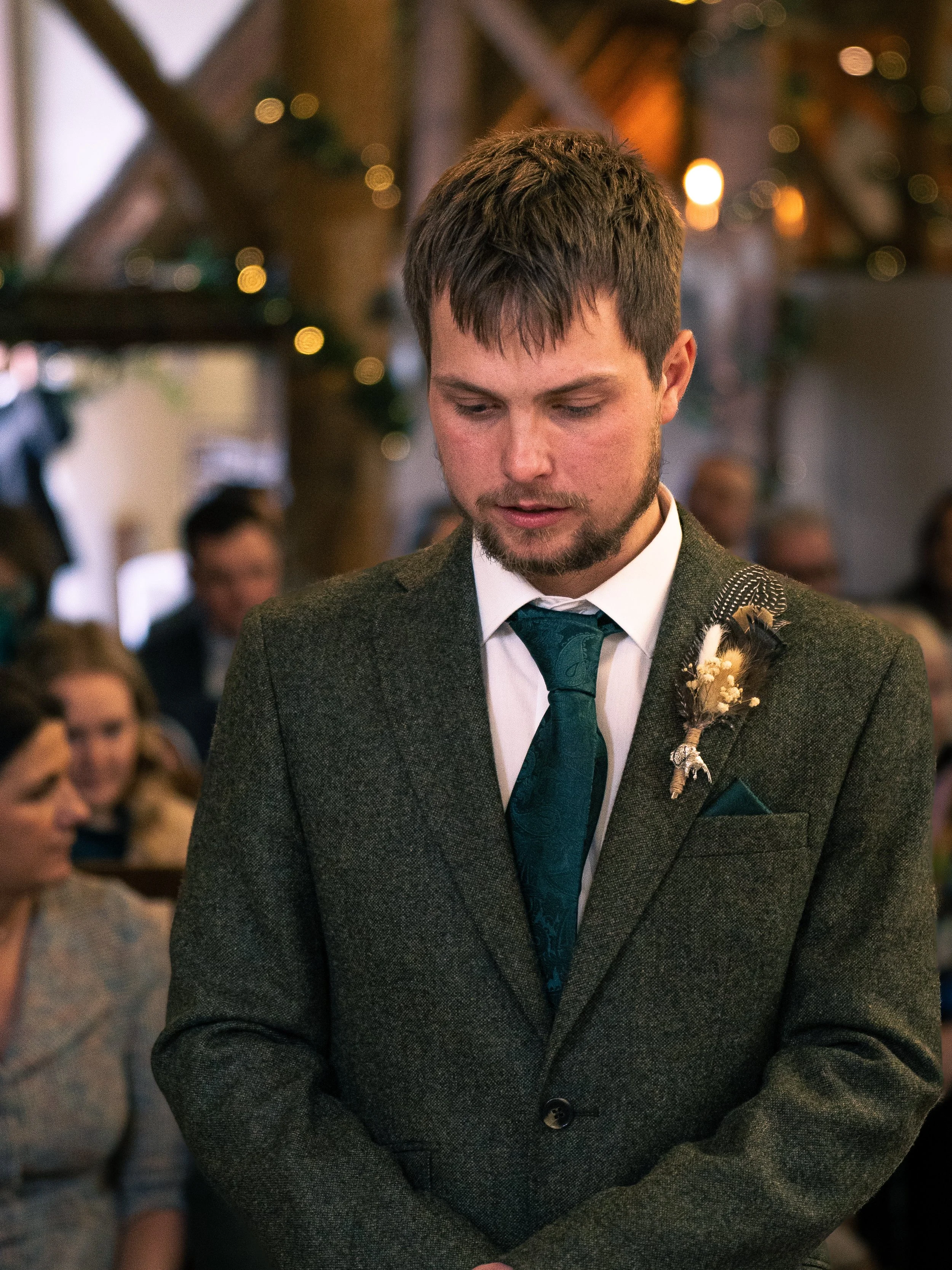 A man in a dark green suit with a boutonnière, looking down during a wedding ceremony in a warmly lit indoor setting.