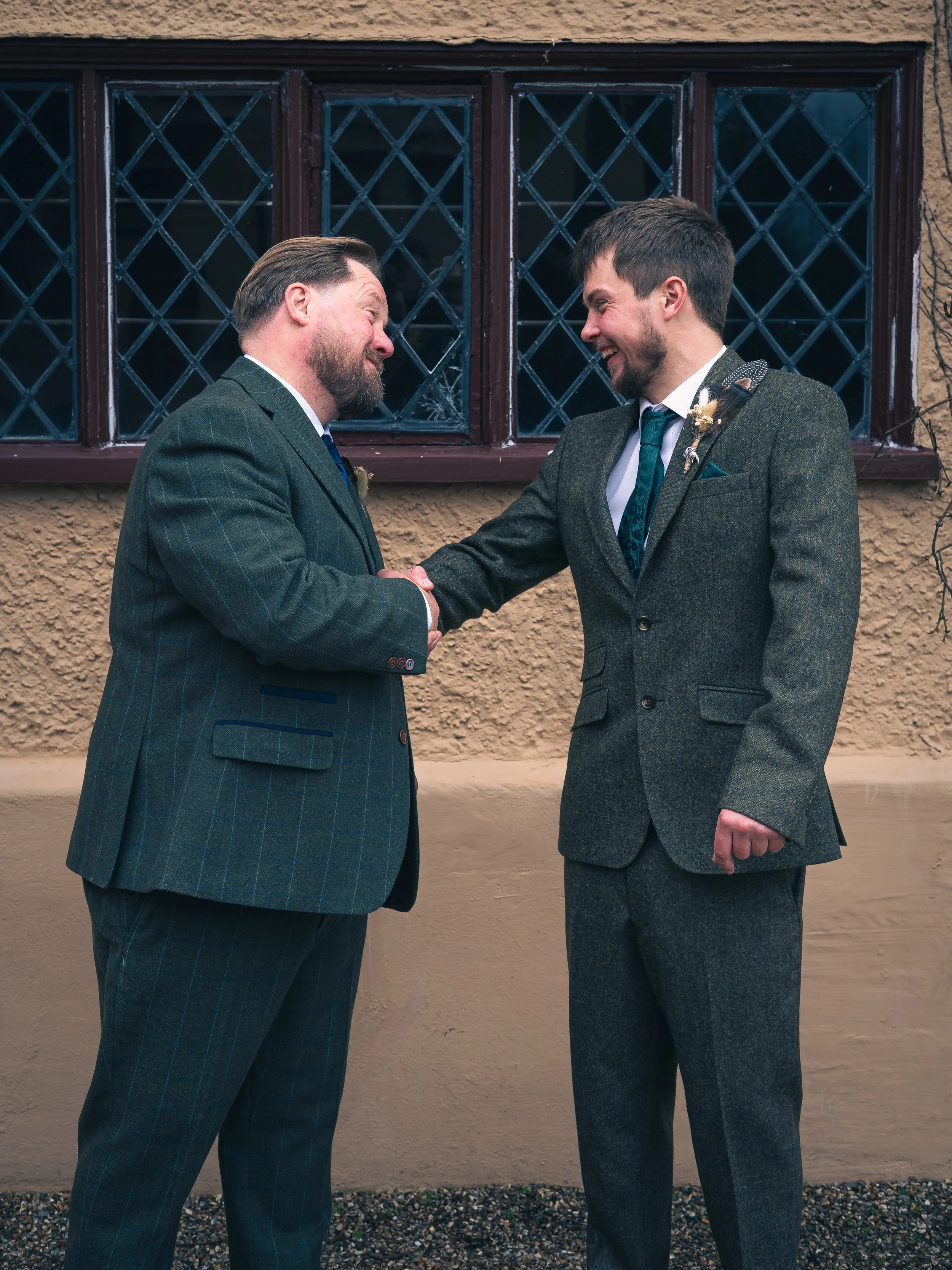 Two men in suits shaking hands and smiling in front of a window with diamond-patterned panes.