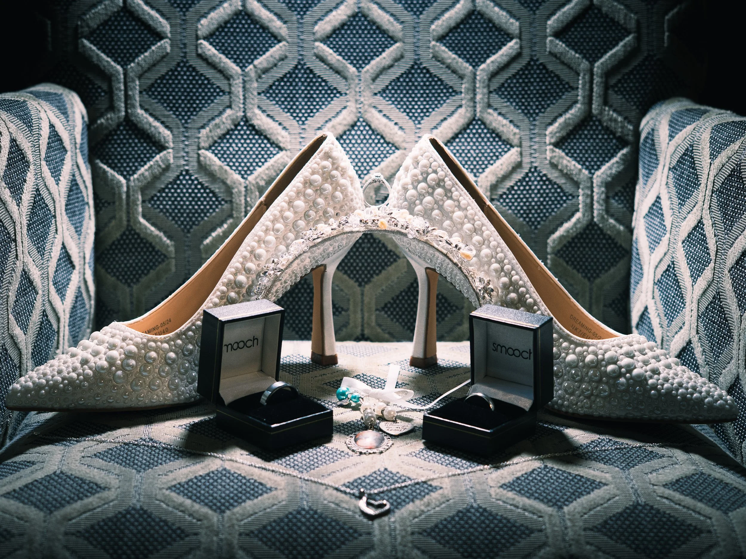 A pair of ornate white high-heeled shoes decorated with pearls and embellishments, displayed on a patterned fabric surface with jewelry and rings in boxes labeled 'mooch' and 'smooch' nearby.