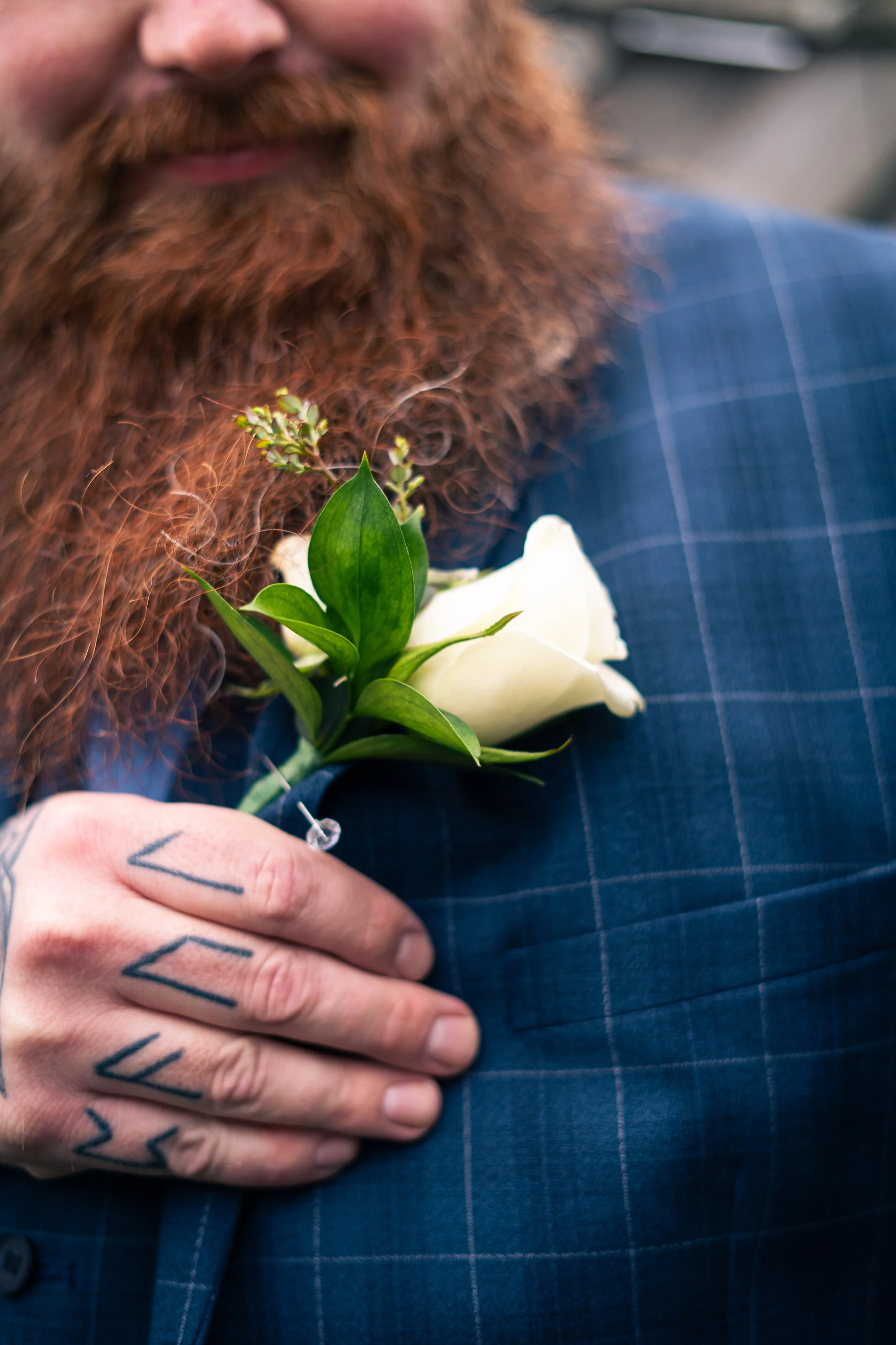 Close-up of a man with a red beard and tattoos on his hand, wearing a blue checkered suit, holding a white rose boutonniere on his chest.