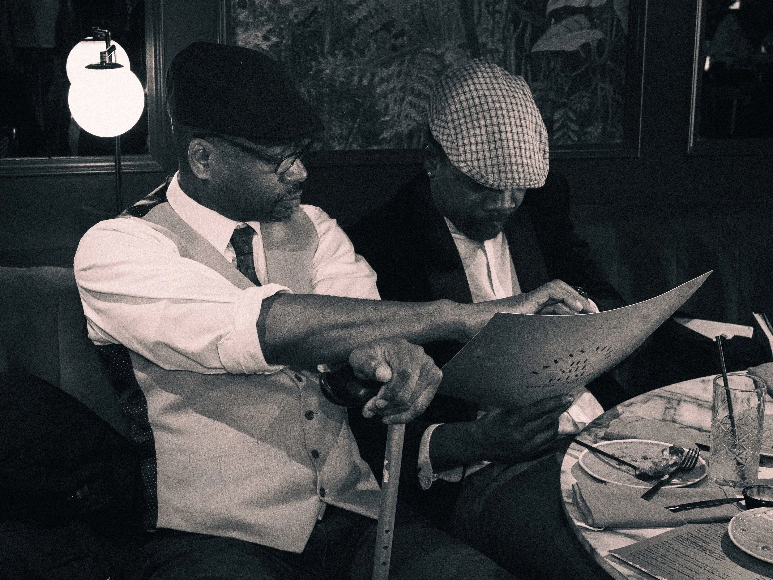 Two men sitting at a table in a restaurant, reviewing a menu together, with plates, glasses, and utensils on the table, and a lamp illuminating the scene.