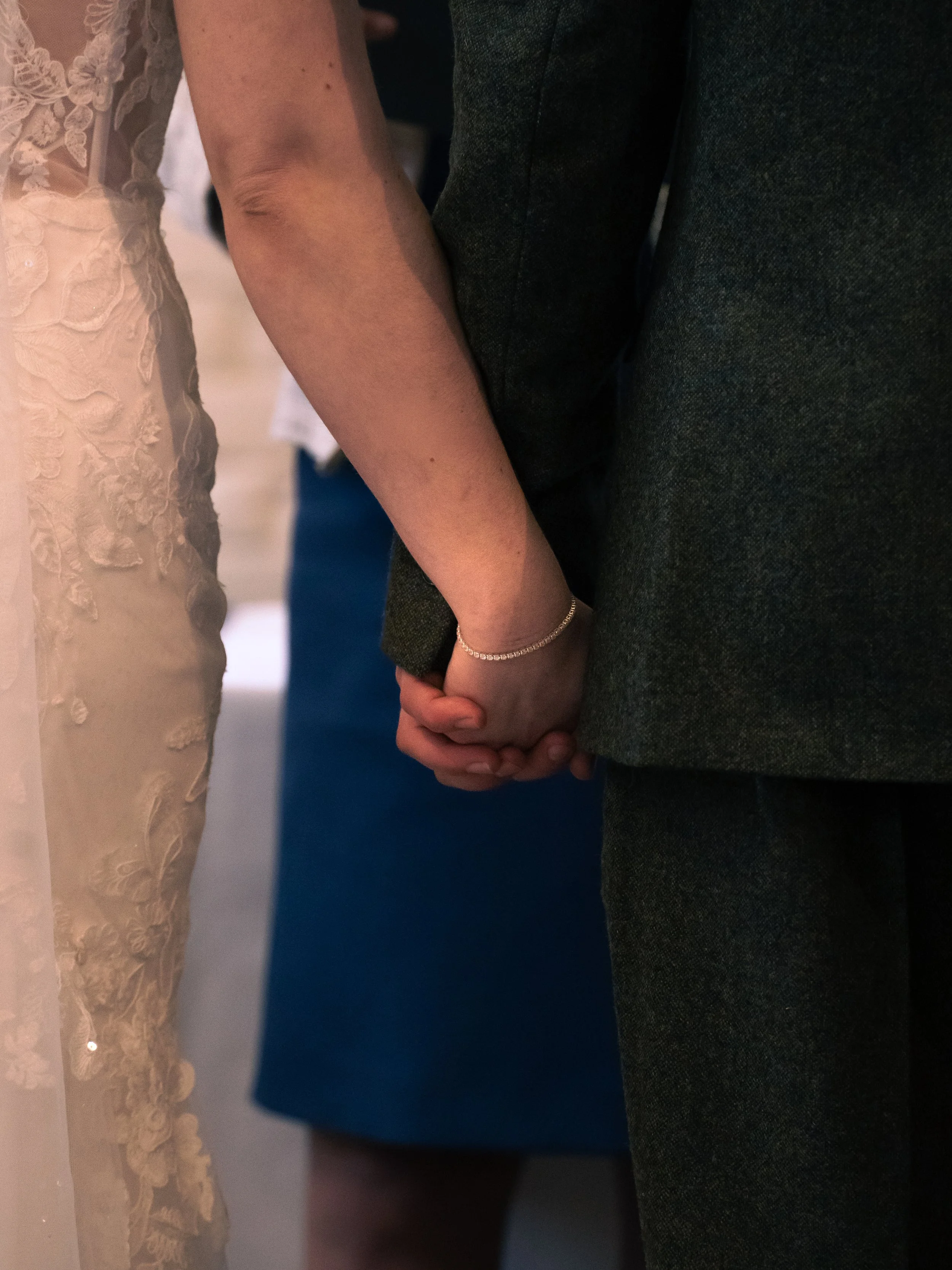 A close-up of a couple holding hands during a wedding, with the bride wearing a lace dress and the groom in a dark suit.