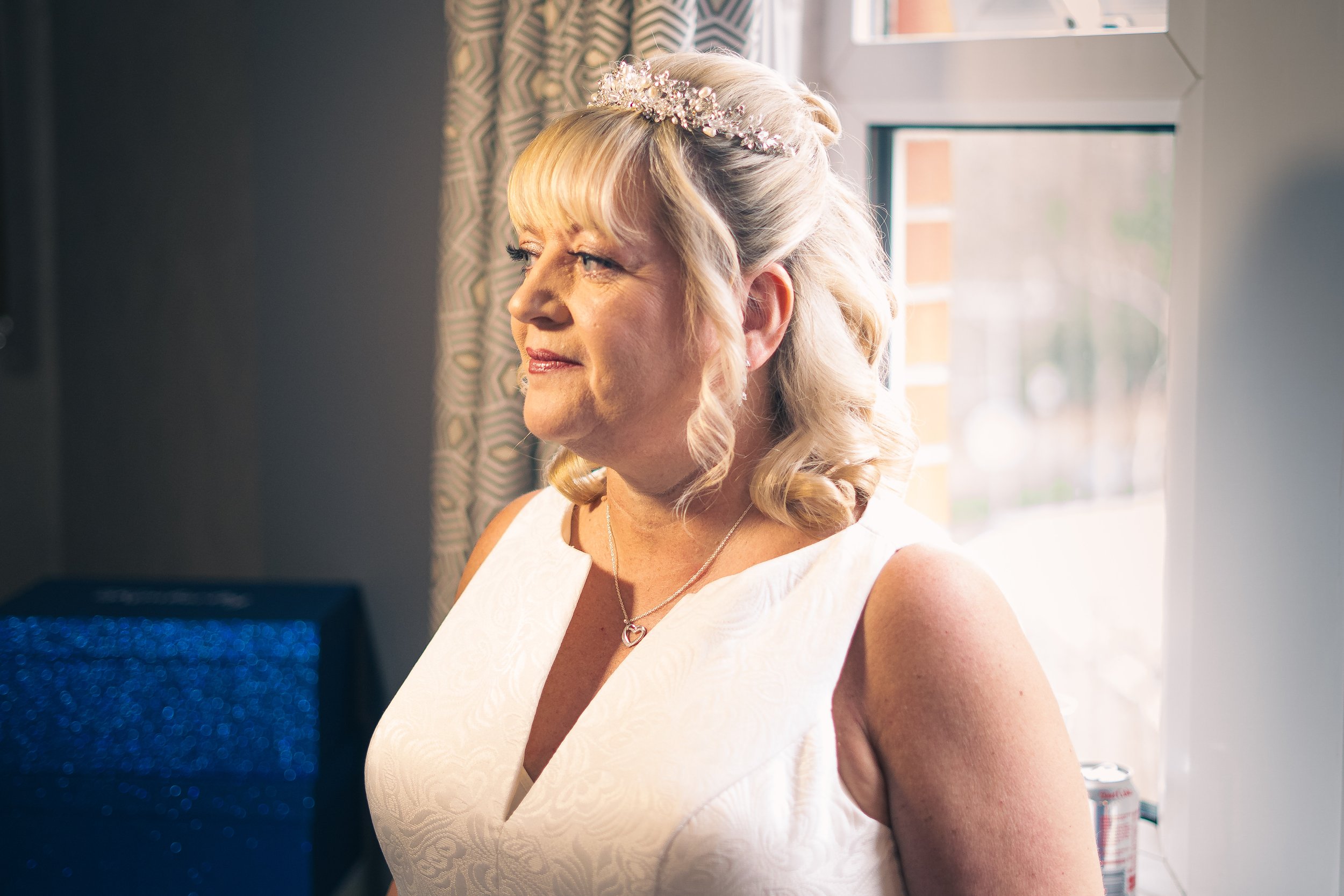 A woman with blonde hair styled in loose curls, wearing a white dress and a floral headband, stands near a window with natural light illuminating her face, looking to the side.