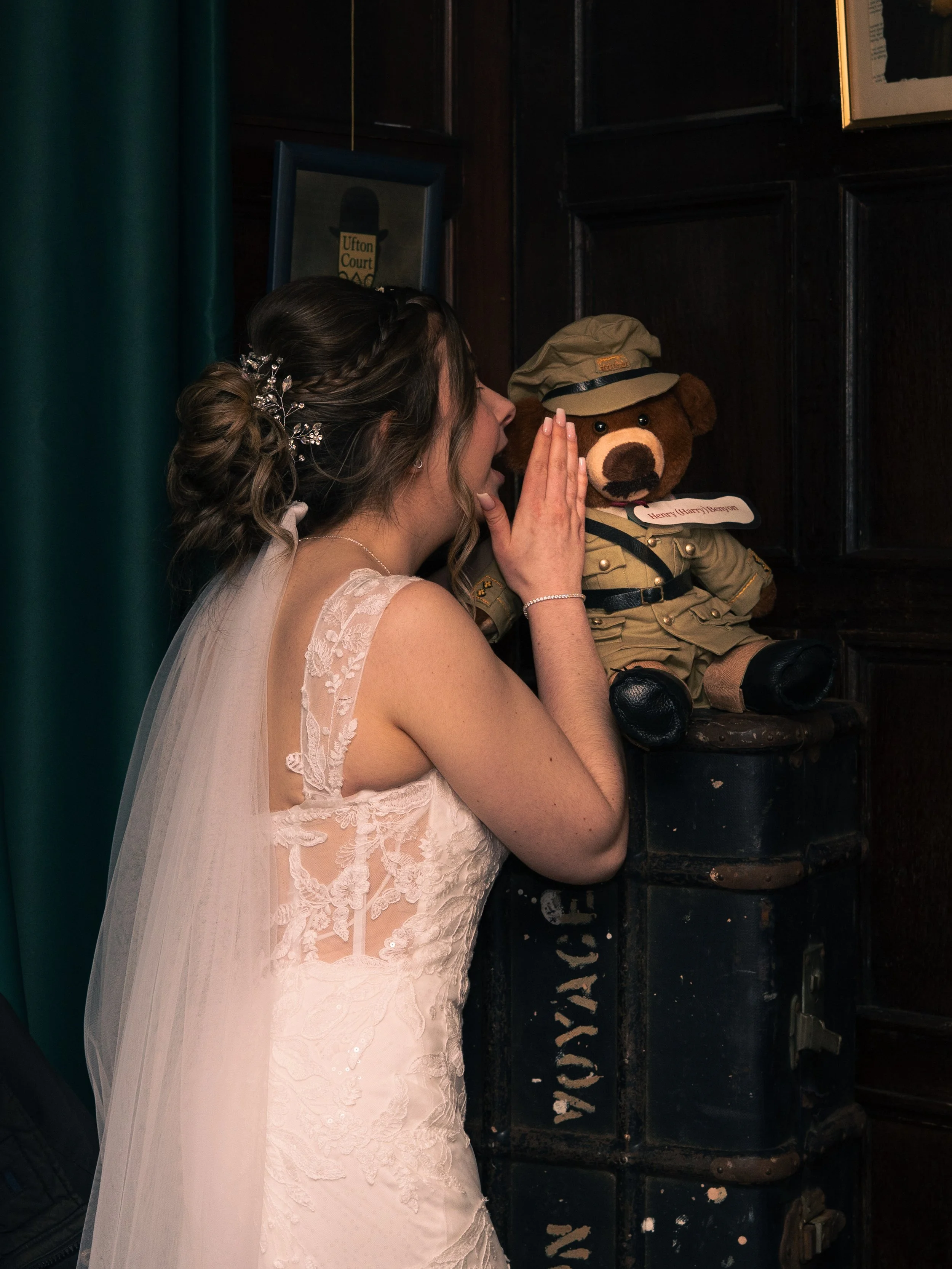A bride in a white lace wedding dress whispers to a stuffed teddy bear dressed as a soldier, positioned on a wooden chest in a dark wood-paneled room.