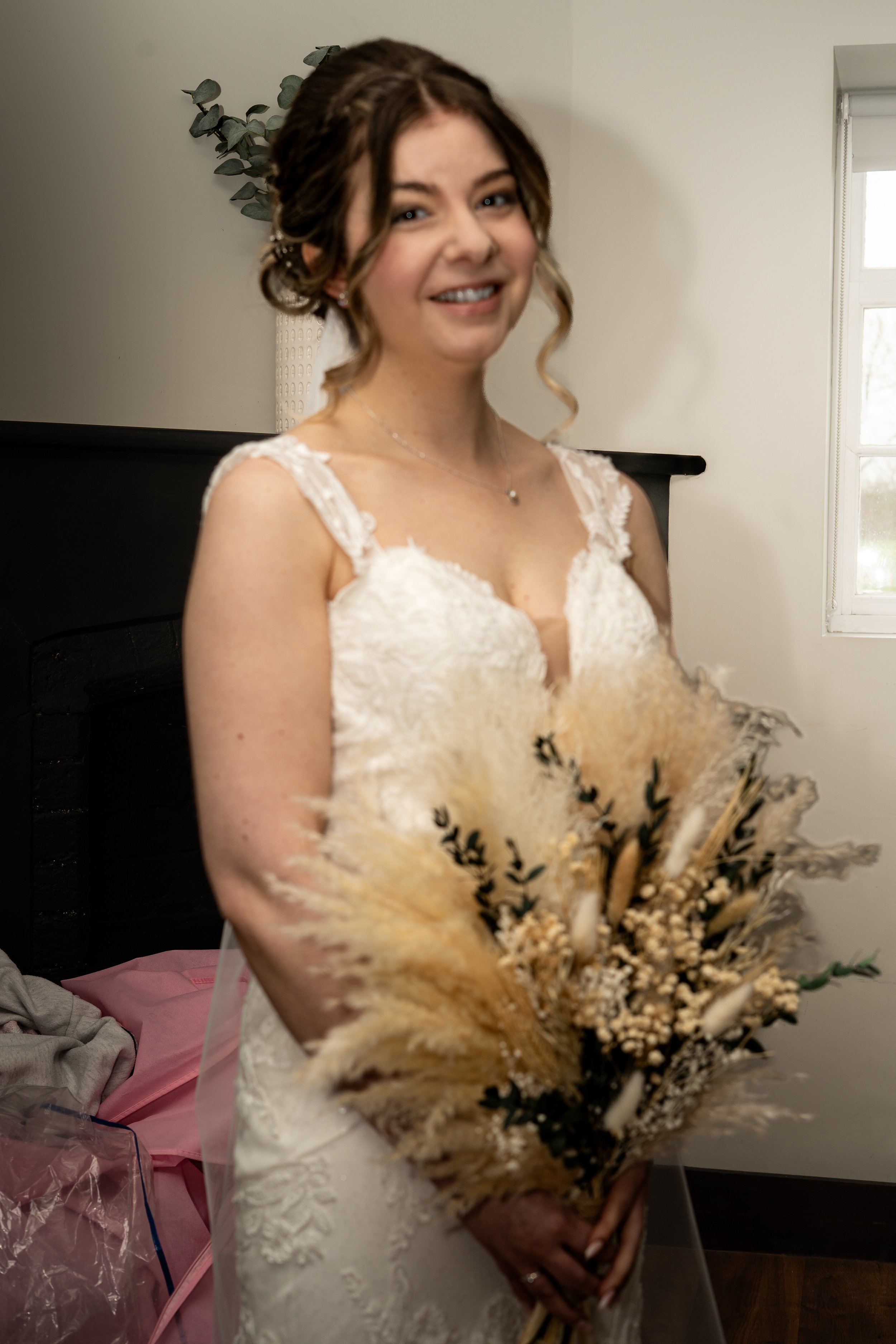 A smiling young woman dressed in a wedding gown holding a bouquet of dried flowers and wheat on a bed in a room with a window and simple decor.