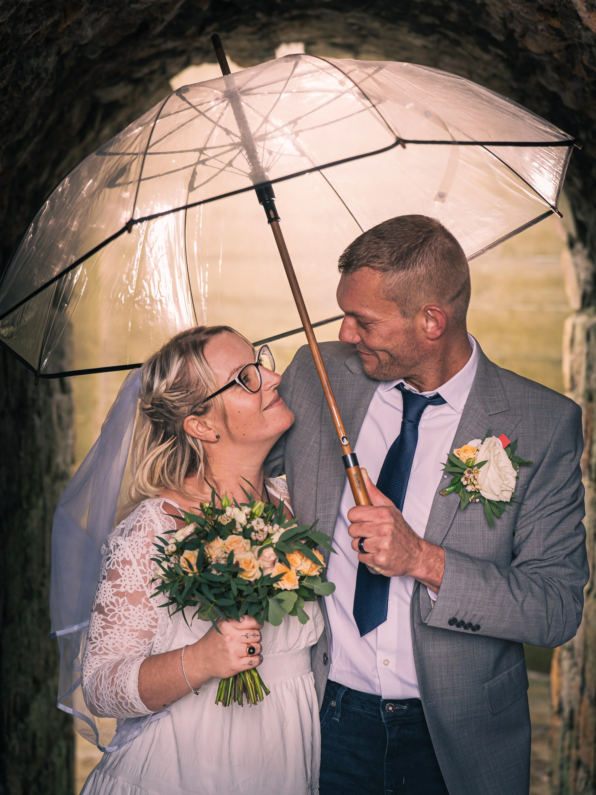 A bride and groom standing under a transparent umbrella outdoors, looking at each other lovingly. The bride is holding a bouquet of flowers, and the groom is dressed in a suit with a boutonniere. They are in front of a natural, wooded background.