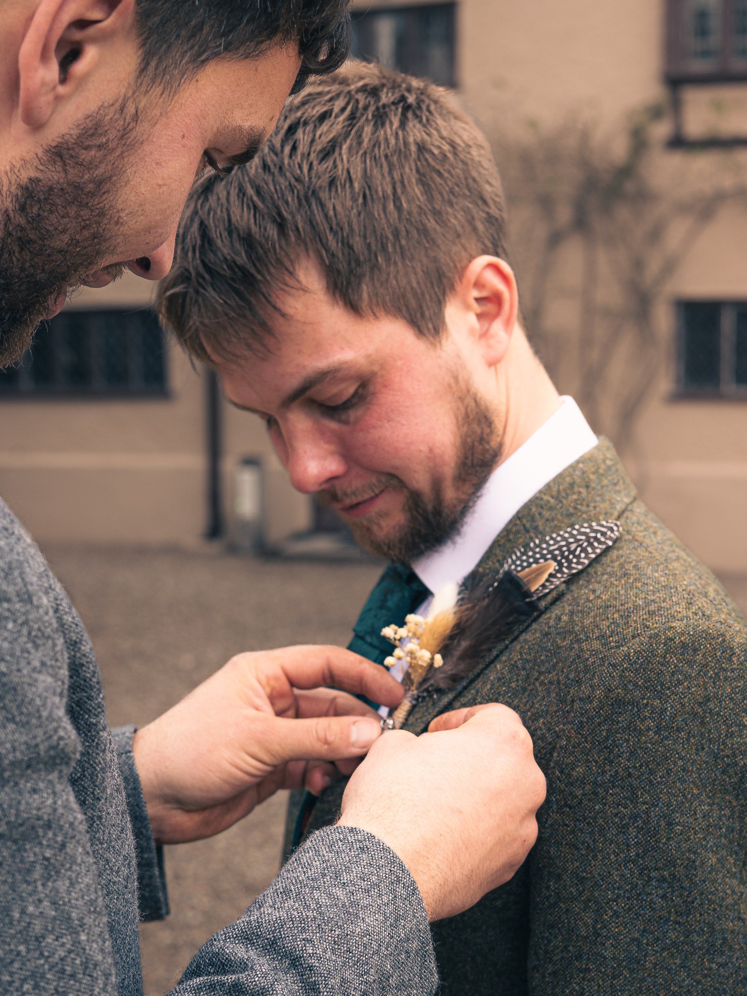 Two men, one pinning a boutonniere onto the other man's suit jacket during an outdoor event.