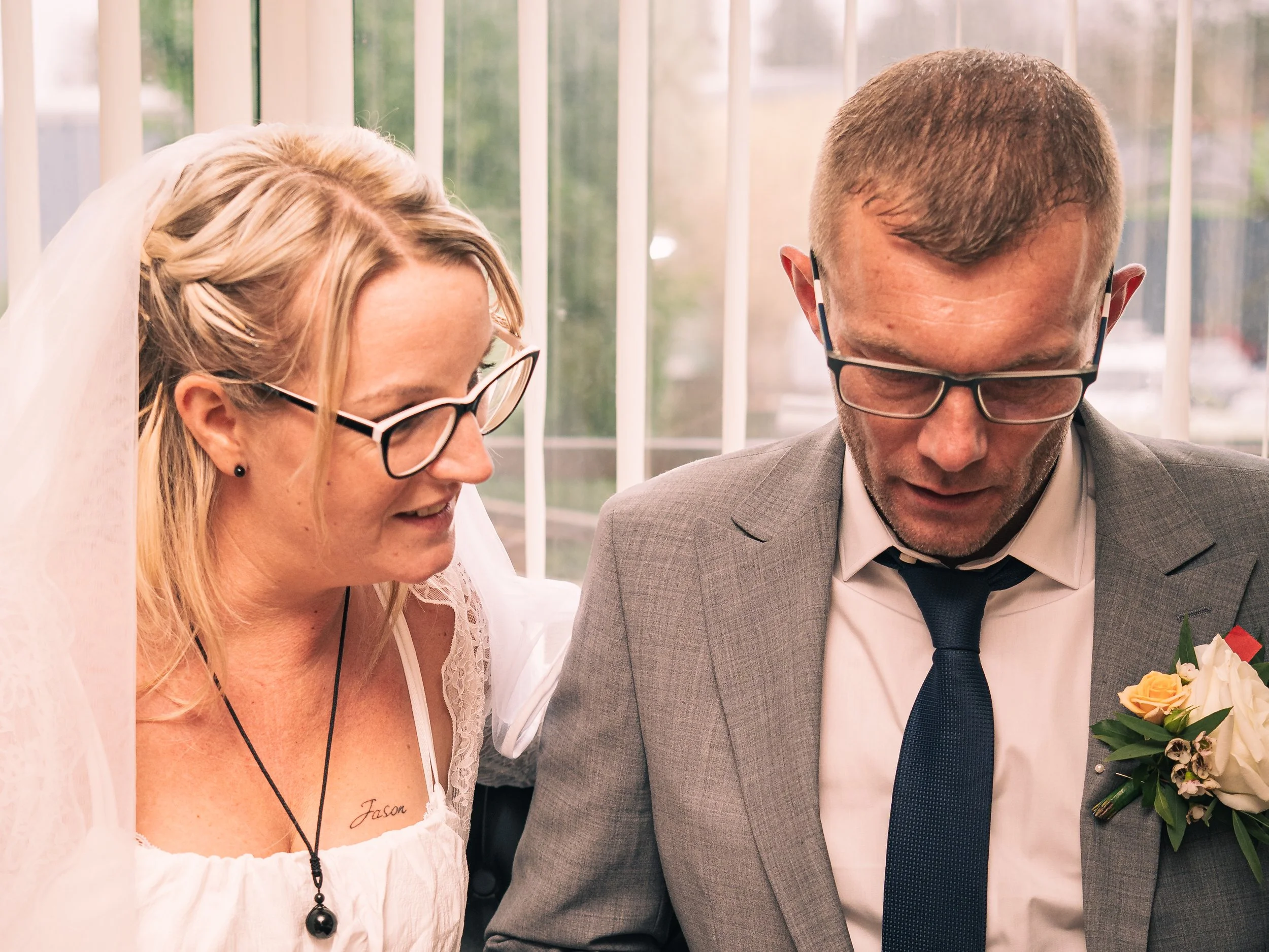 A bride with glasses and a tattoo on her chest is smiling, standing next to a man in a gray suit with a tie, glasses, and a boutonniere, both leaning in close together in front of a window.