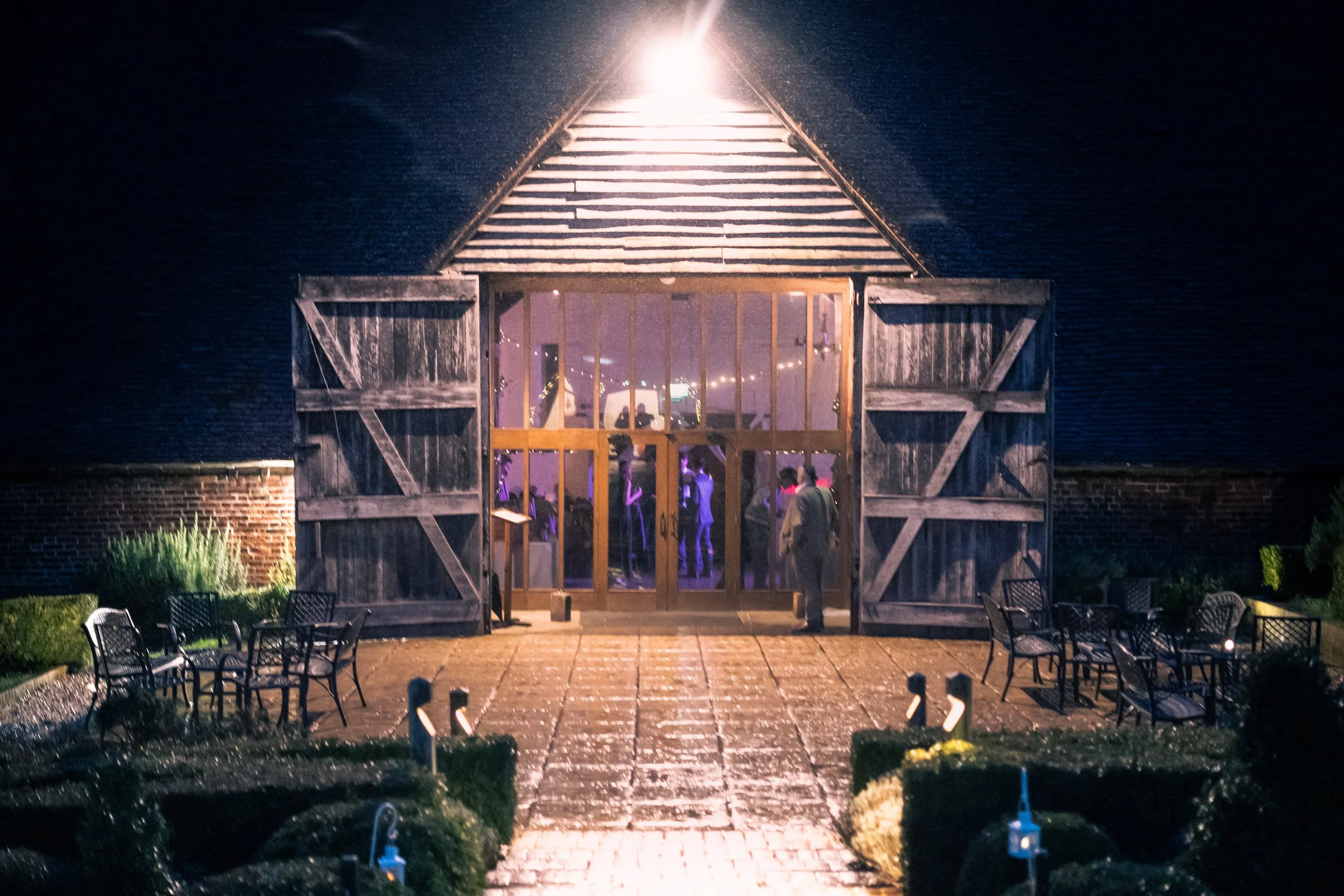 A barn converted into an event space with open doors revealing guests inside, illuminated with purple lighting. The exterior features a paved walkway and outdoor seating on either side, with greenery and hedges surrounding the area.