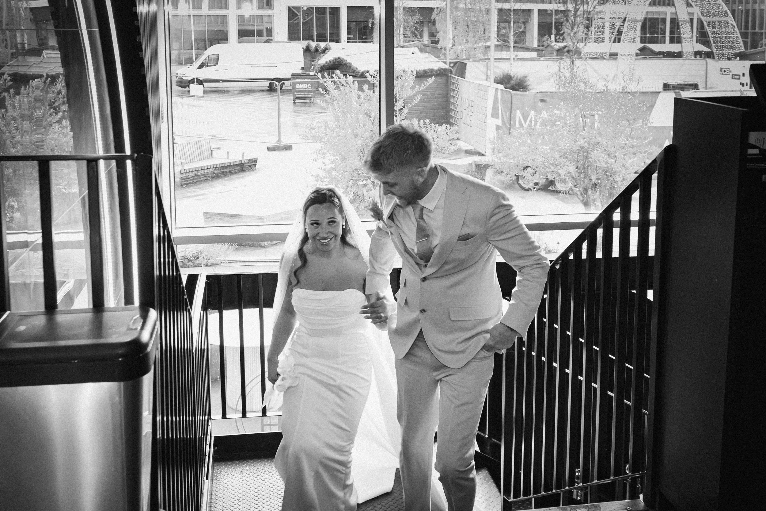 A bride and groom descending stairs at their wedding celebration, smiling inside a building with large windows showing the street outside.