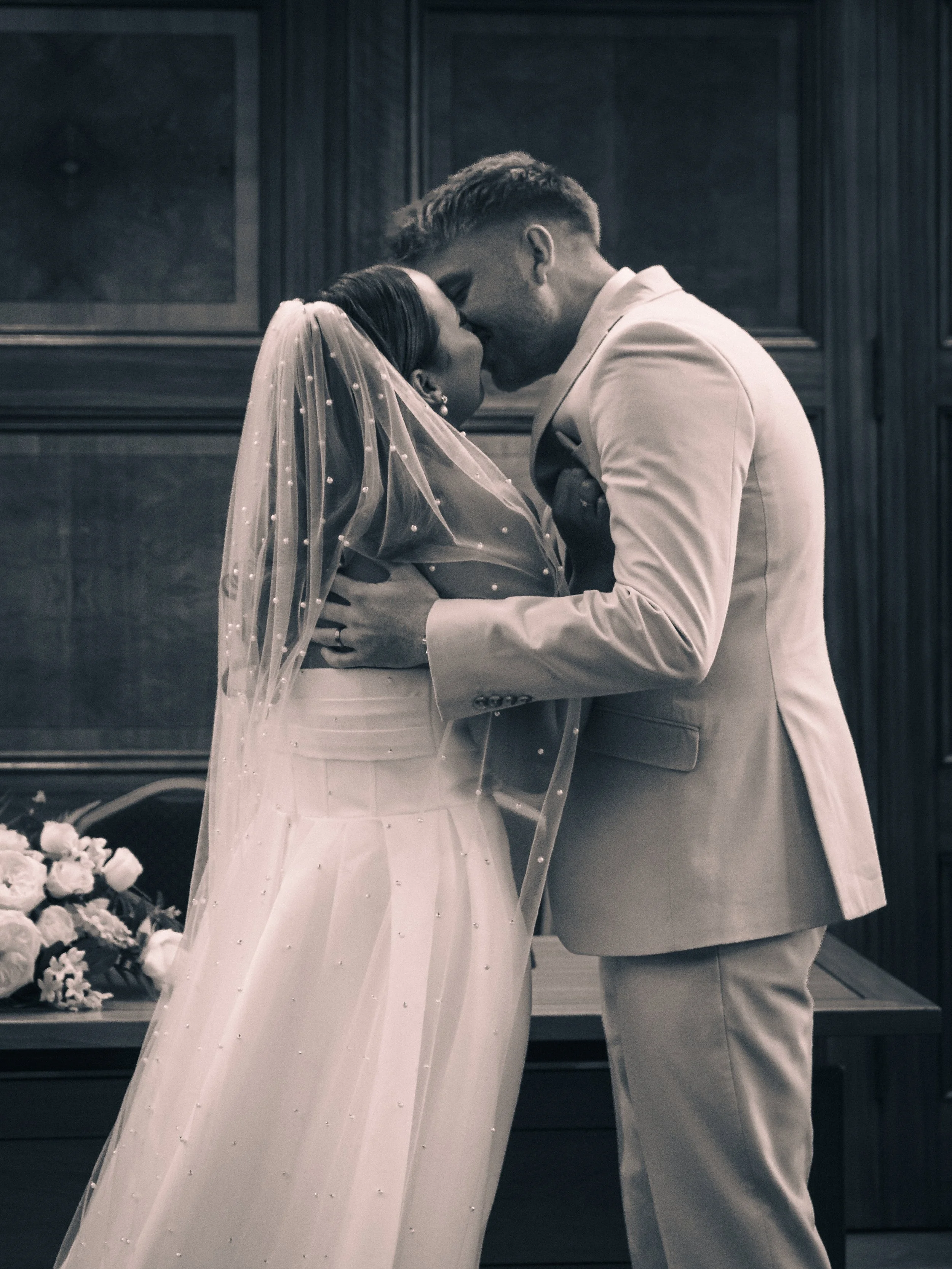 Black and white photo of a bride and groom sharing a kiss during their wedding ceremony, with the bride wearing a veil and gown, and the groom in a suit.