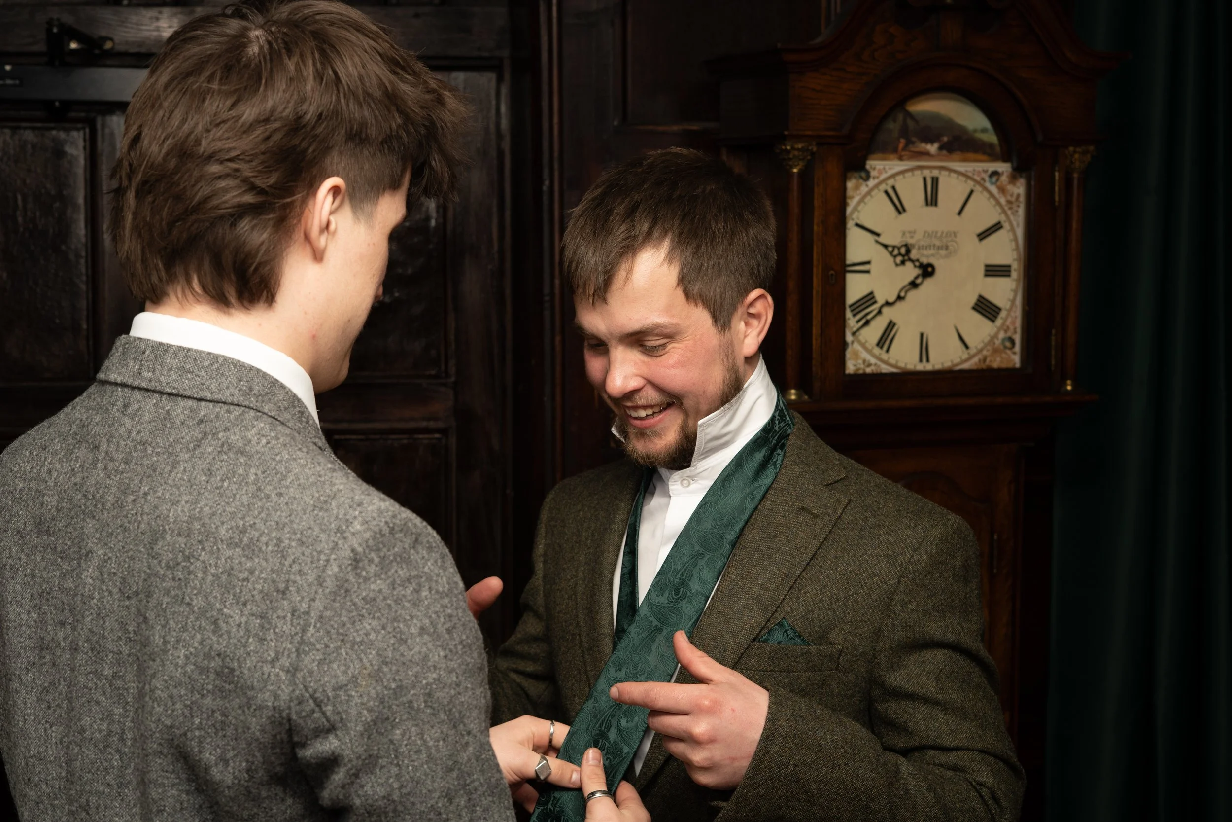 Two men dressed in suits, one smiling, exchange a medal or decoration in a formal setting with a dark wooden clock in the background.