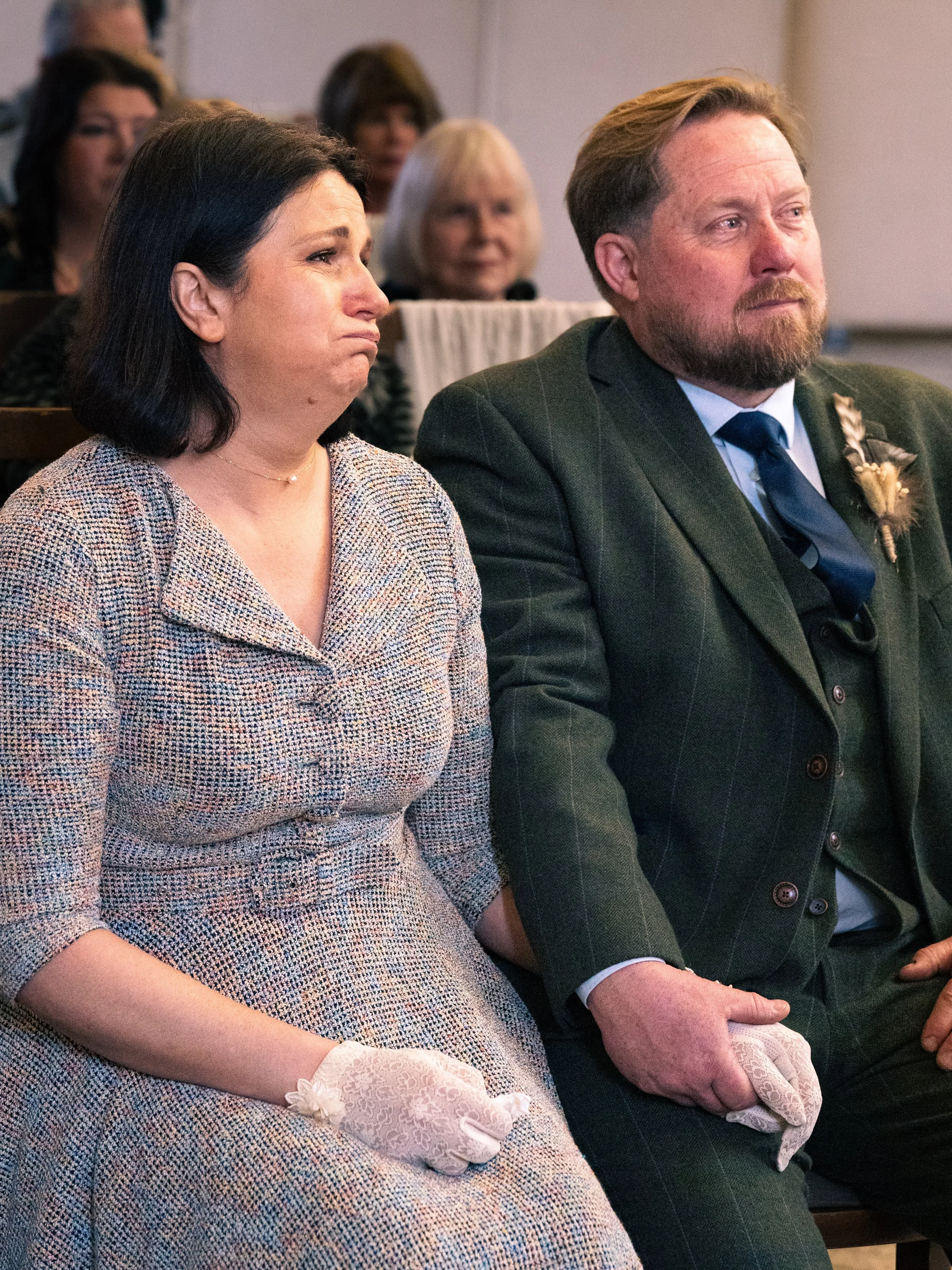 A woman and man sitting side by side during a formal event, both with somber expressions. The woman has shoulder-length dark hair and is wearing a patterned dress with white lace gloves. The man has a beard and is dressed in a dark suit with a blue t