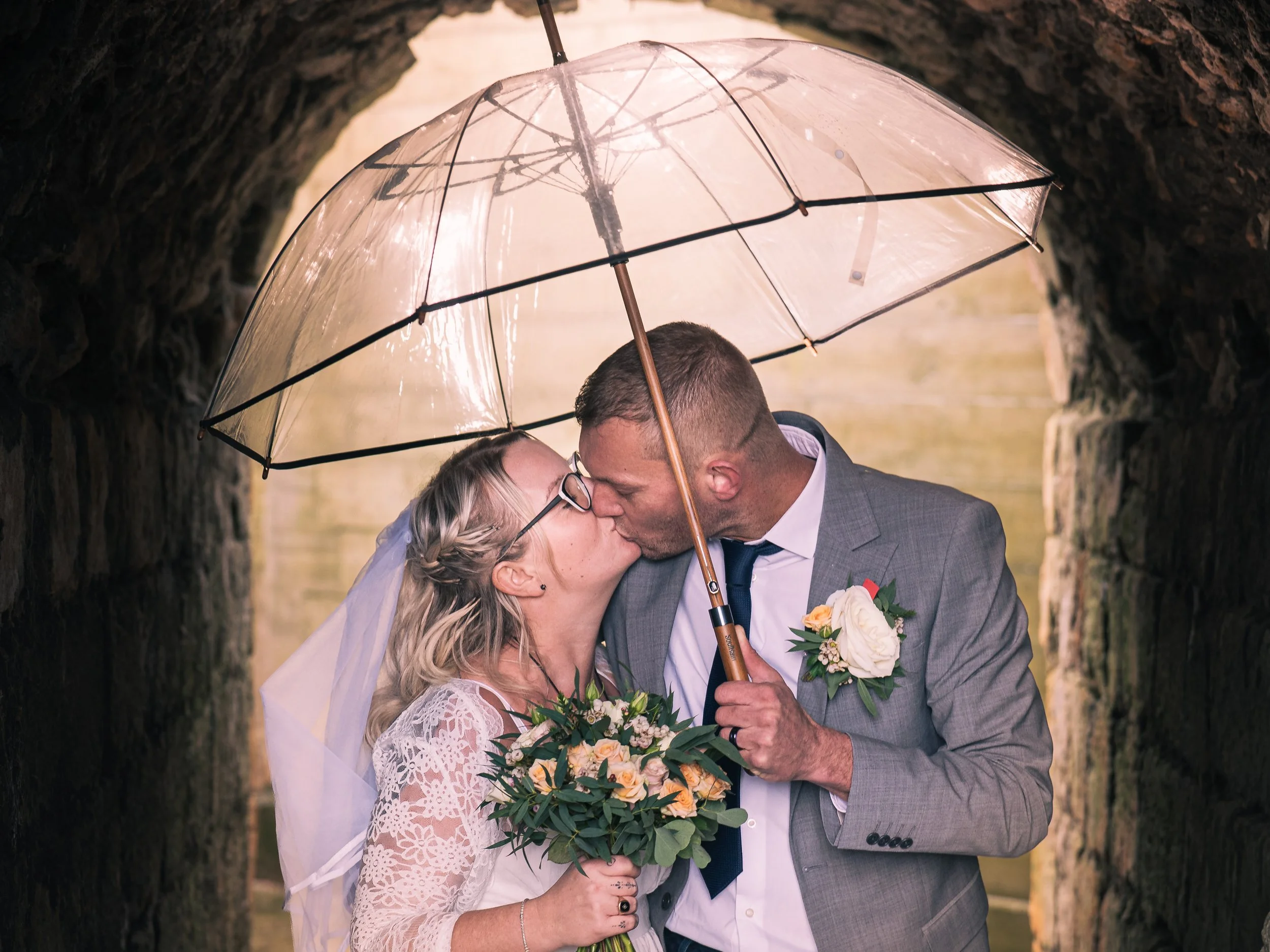 A bride and groom kissing under a transparent umbrella in a stone archway, the bride holding a bouquet of flowers and the groom in a gray suit with a boutonniere.