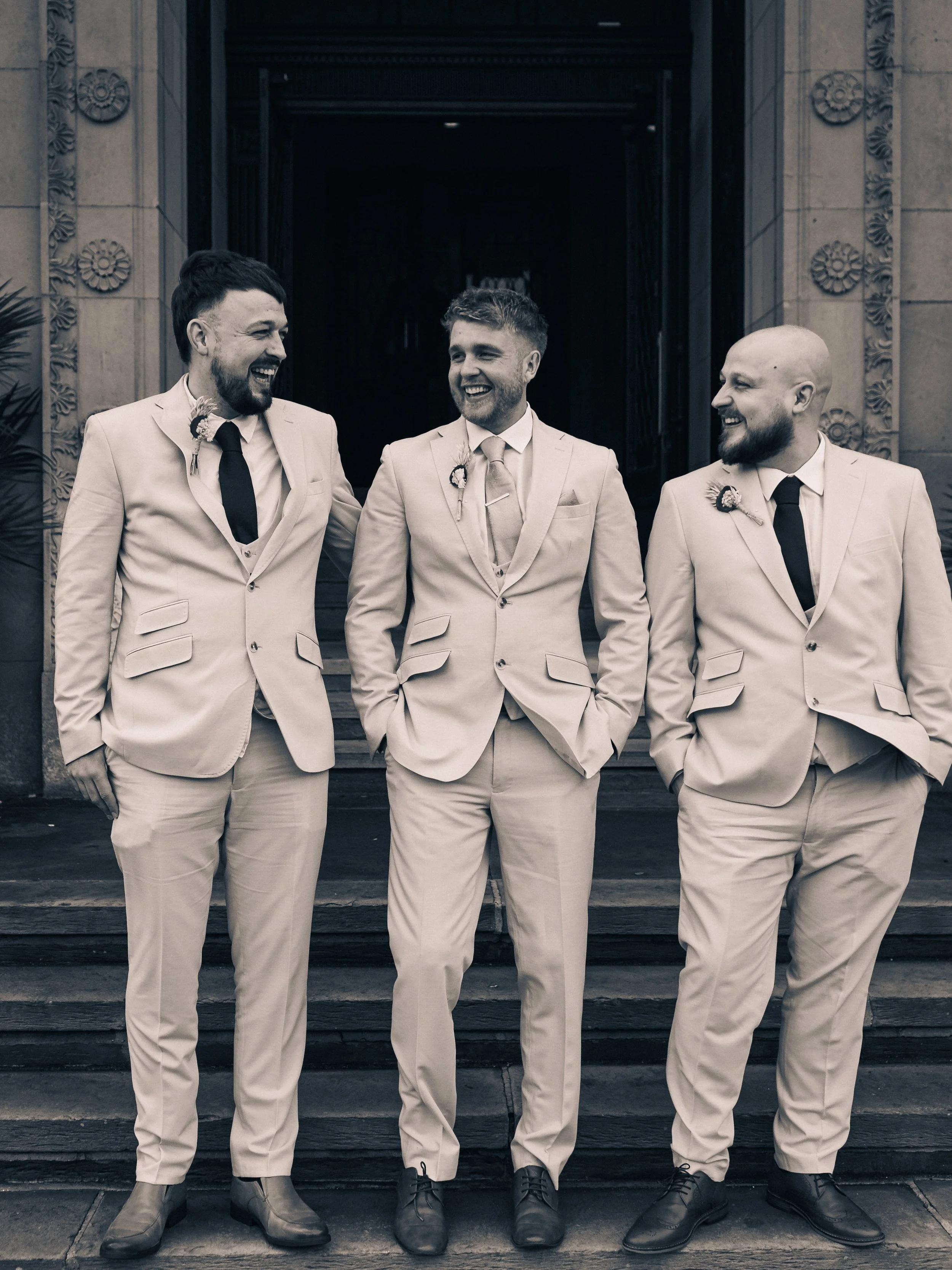 Three men in light-colored suits standing on steps outside a building, smiling and chatting with each other.