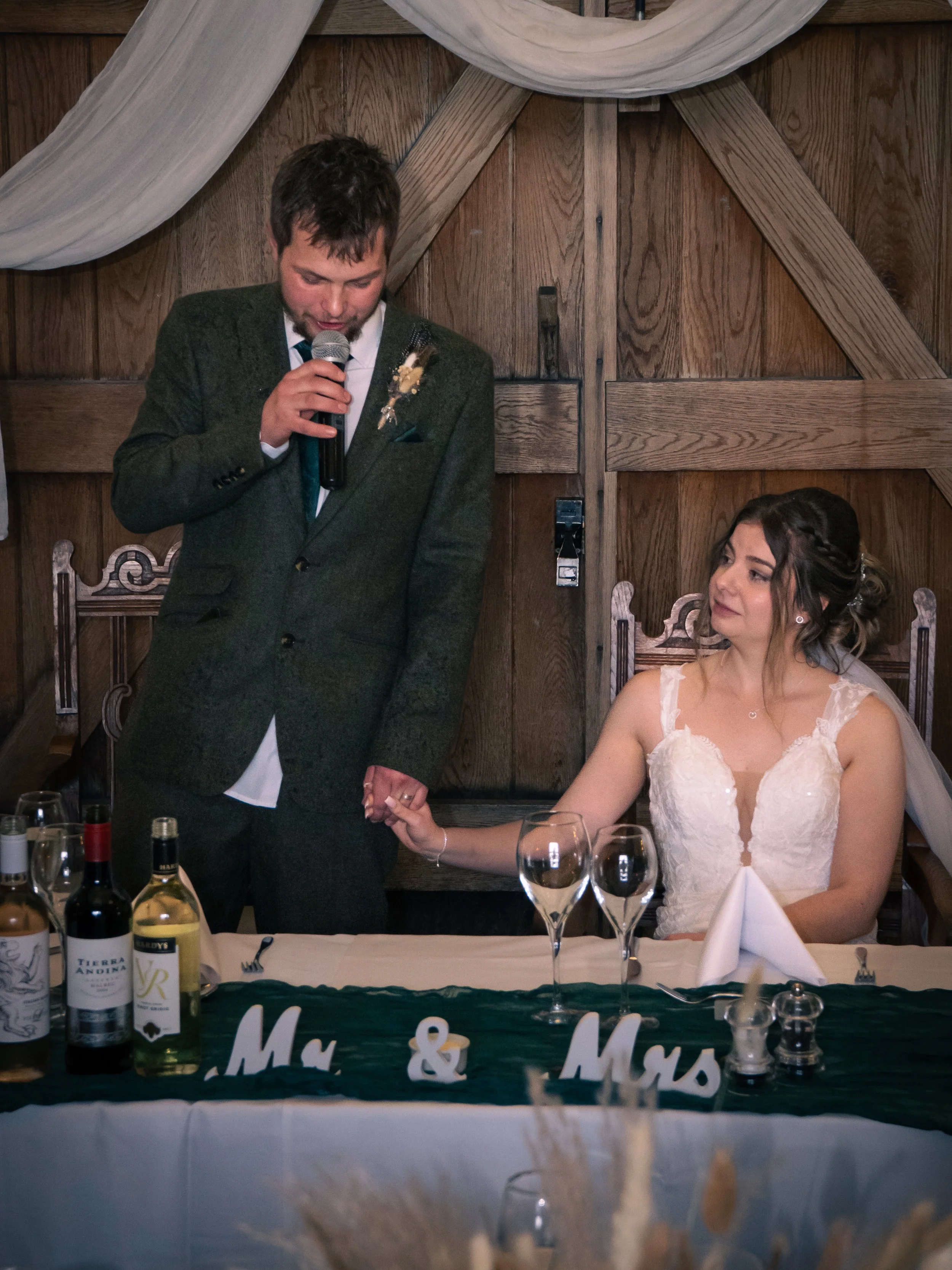 A groom in a dark suit and a bride in a white lace wedding dress holding hands during a wedding reception, with the groom speaking into a microphone and the bride seated at a table decorated with 'Mr & Mrs' signs and wine bottles.