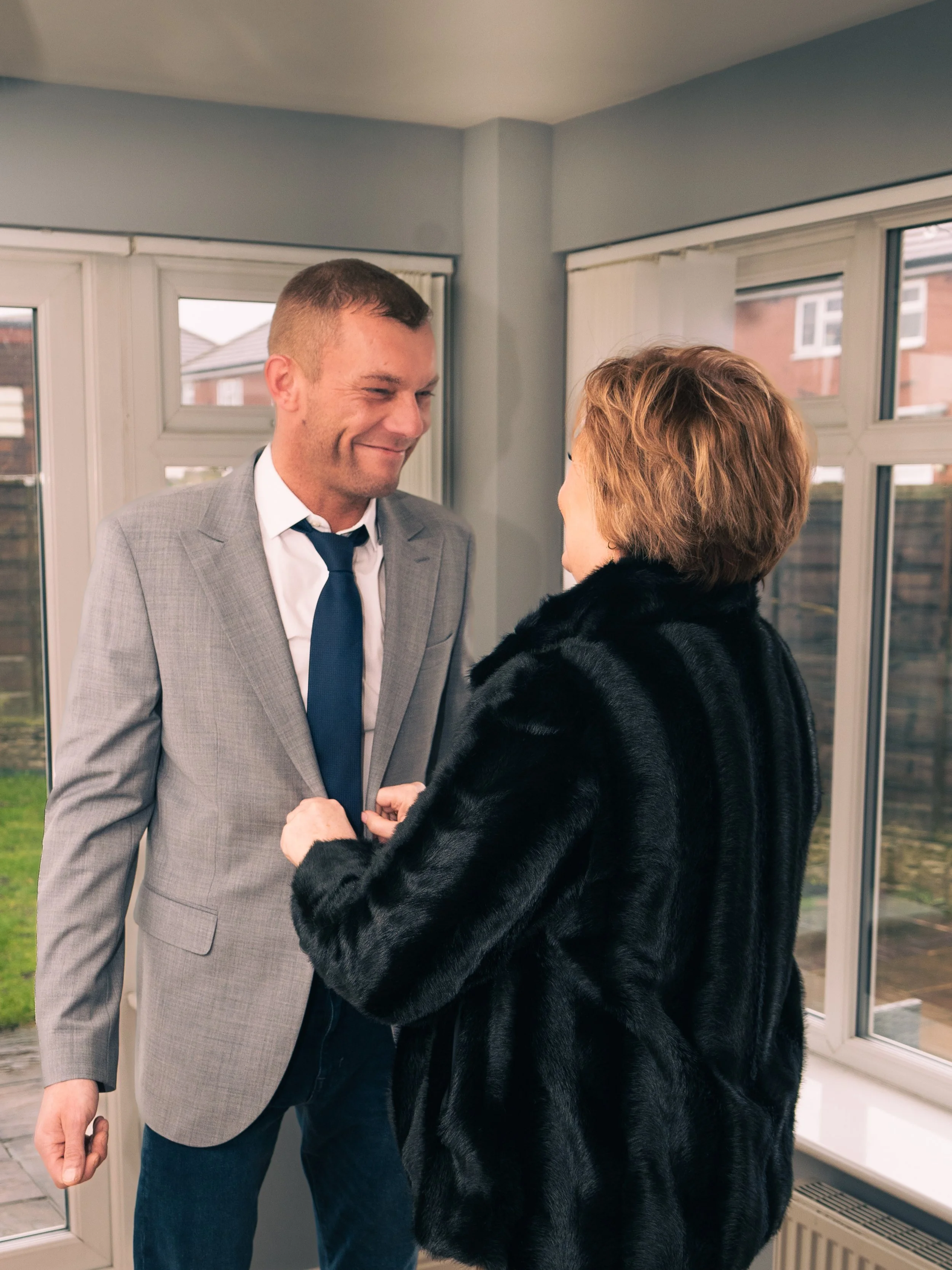 A man in a gray suit and a woman in a black fur coat are smiling and holding hands in a room with large windows showing the outside.
