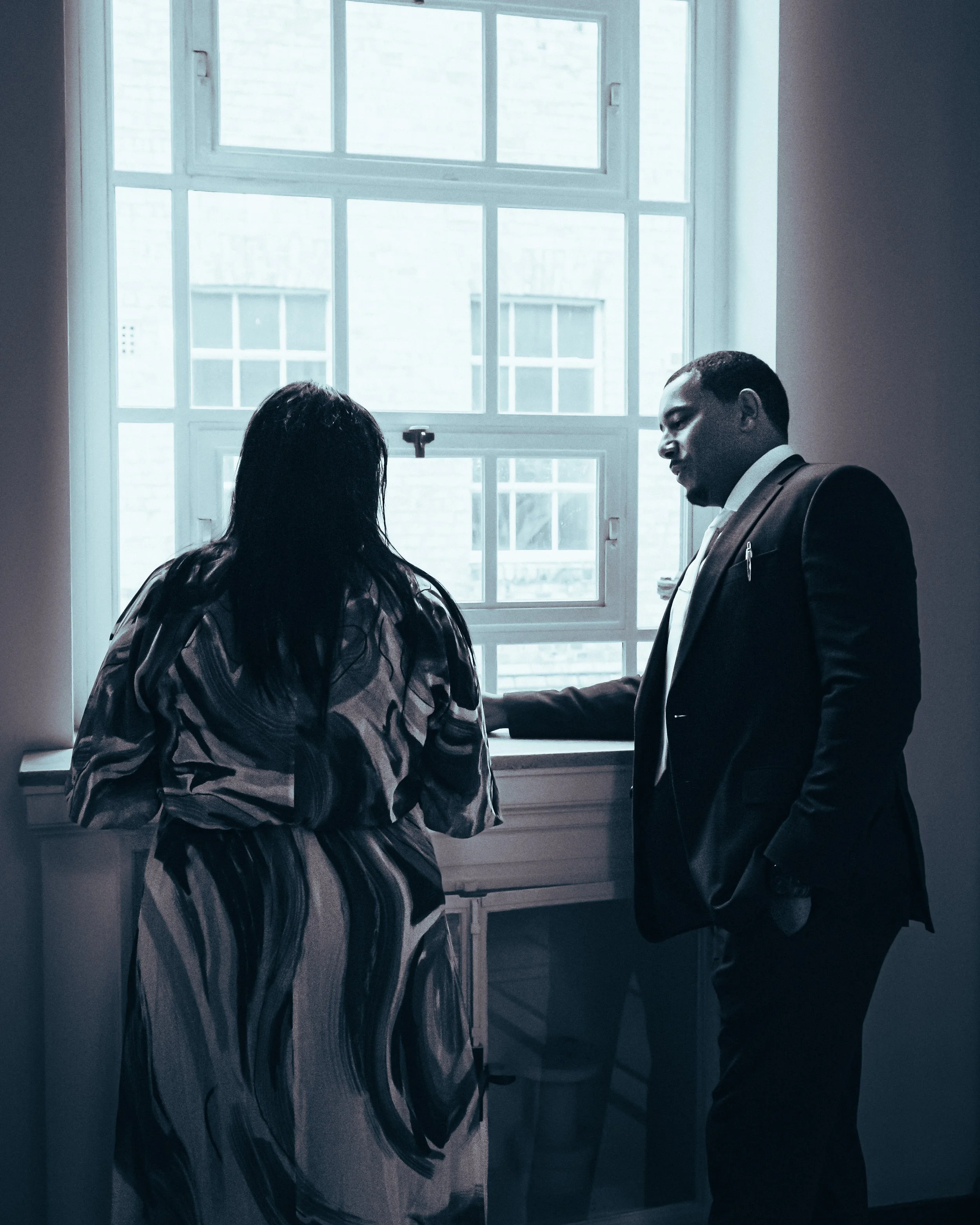 A man and woman standing near a window, engaged in a conversation indoors during the daytime.