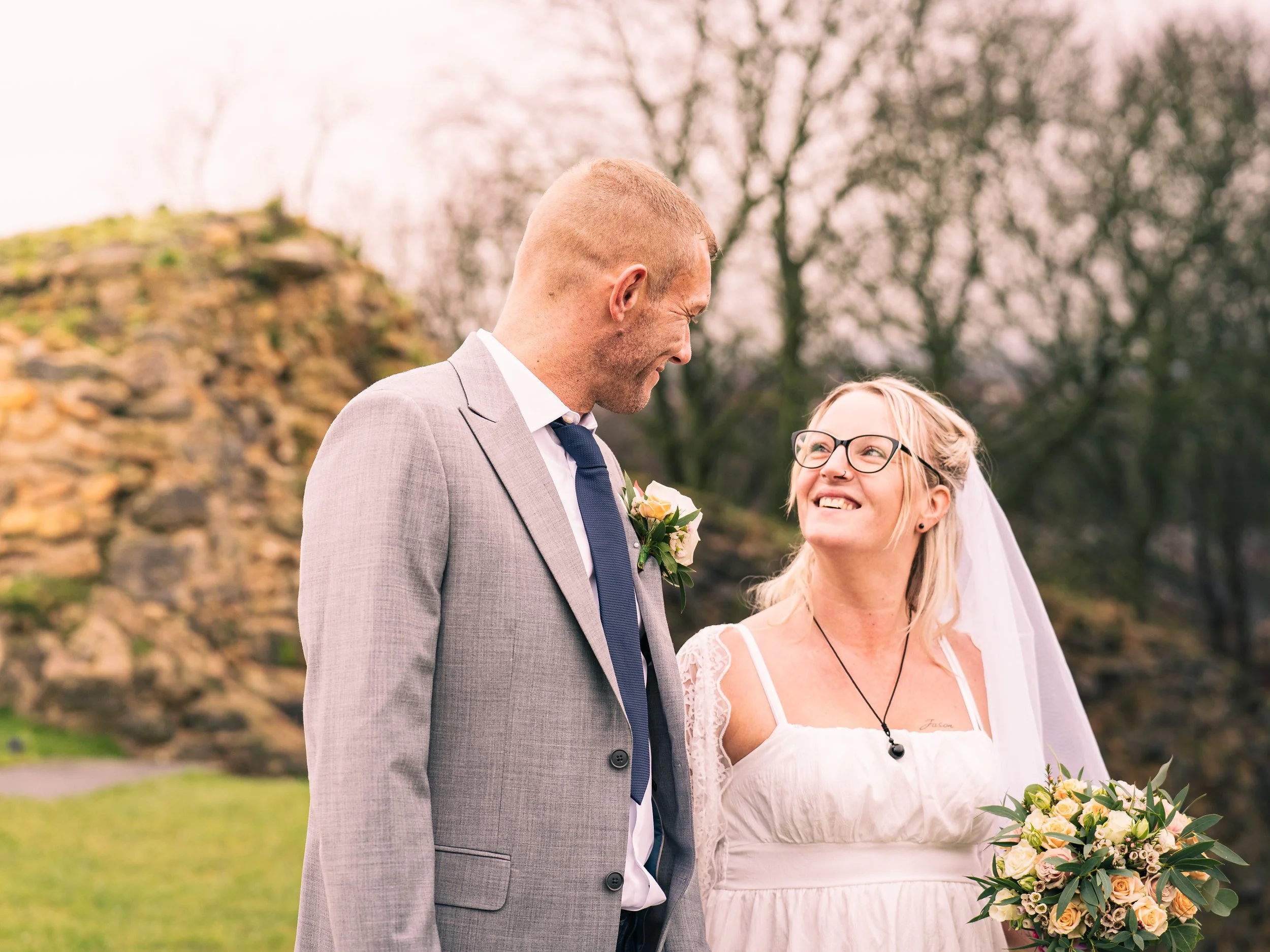 A bride and groom standing outdoors, smiling at each other, with trees and a stone structure in the background.