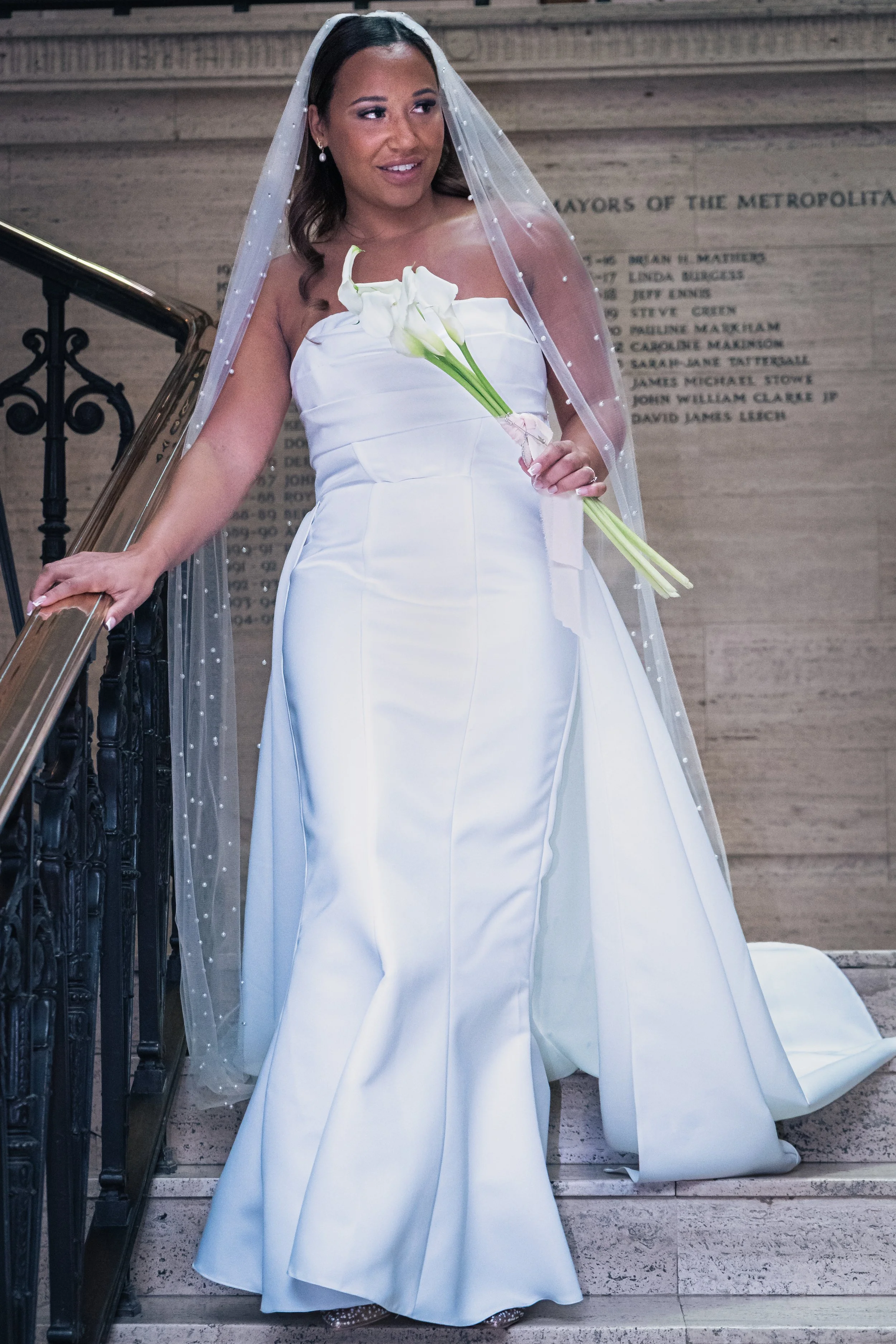 A woman in a wedding dress holding a bouquet of white calla lilies on a staircase.