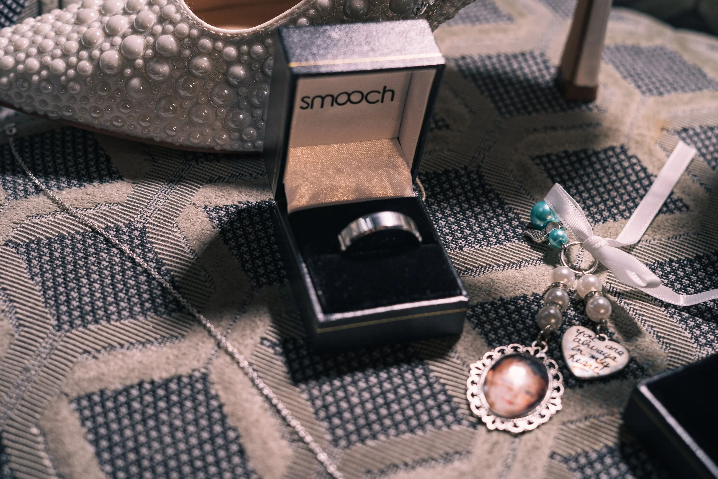 A jewelry collection on a patterned fabric surface, including a pearl embellished clutch, a silver ring in an open Smooch box, a pearl necklace with a white ribbon, and a necklace with a heart-shaped charm with handwritten message.