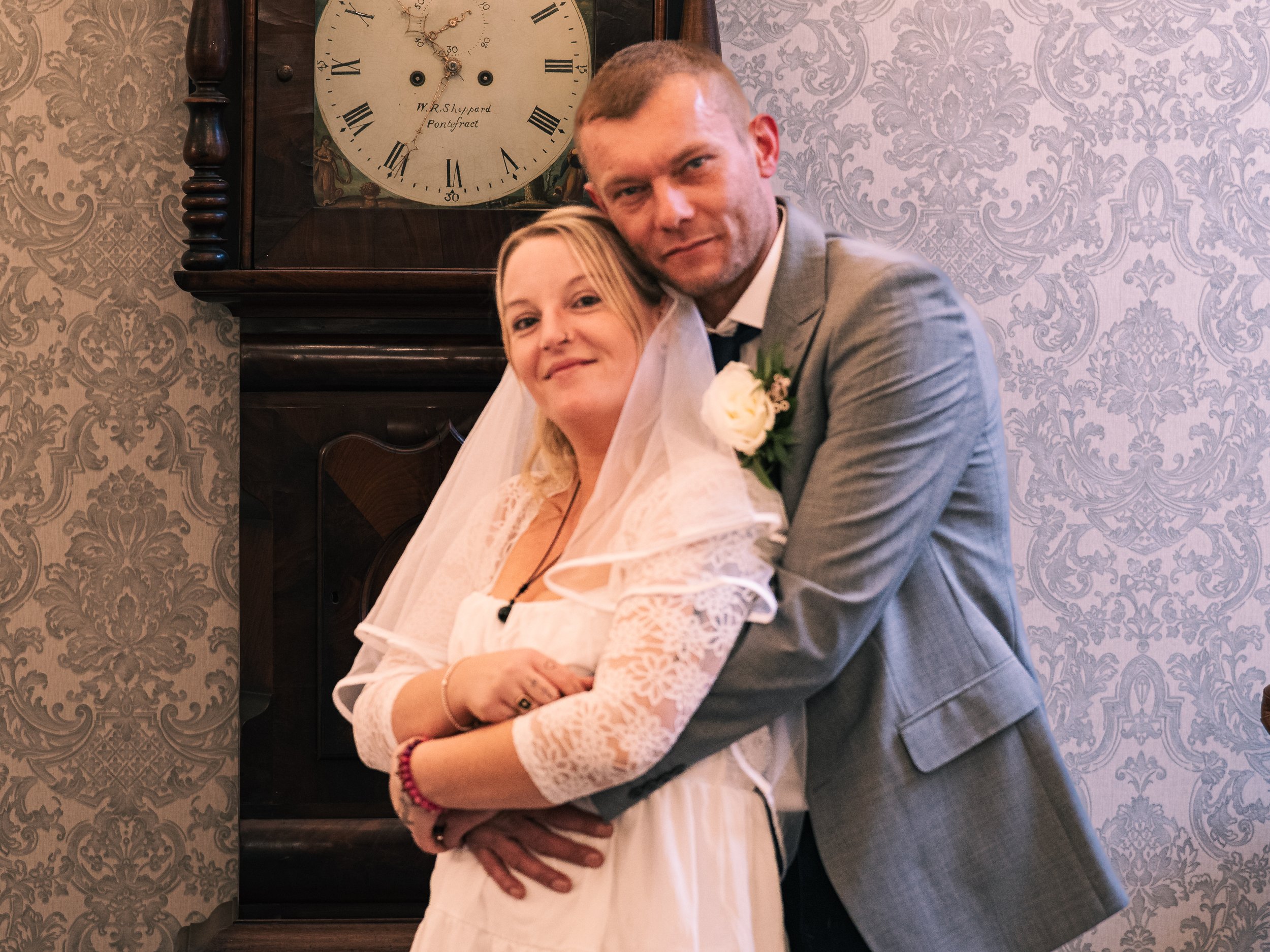 A bride and groom on their wedding day, standing close together indoors with ornate wallpaper and a large antique clock in the background.