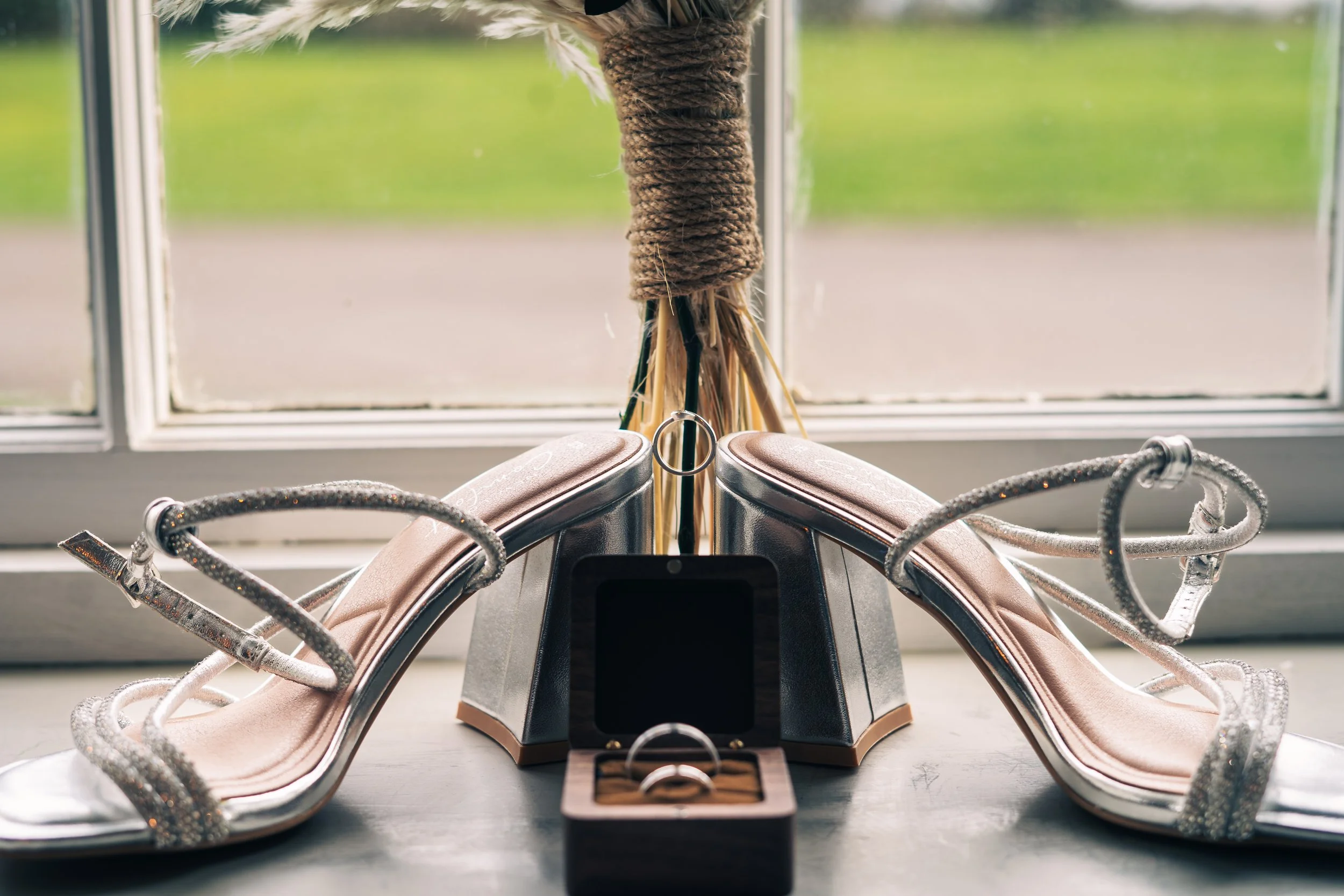 A pair of elegant high-heeled shoes with glittering straps, a ring in a wooden box, and a silver ring displayed in a small box, all positioned on a windowsill with a blurred outdoor view in the background.