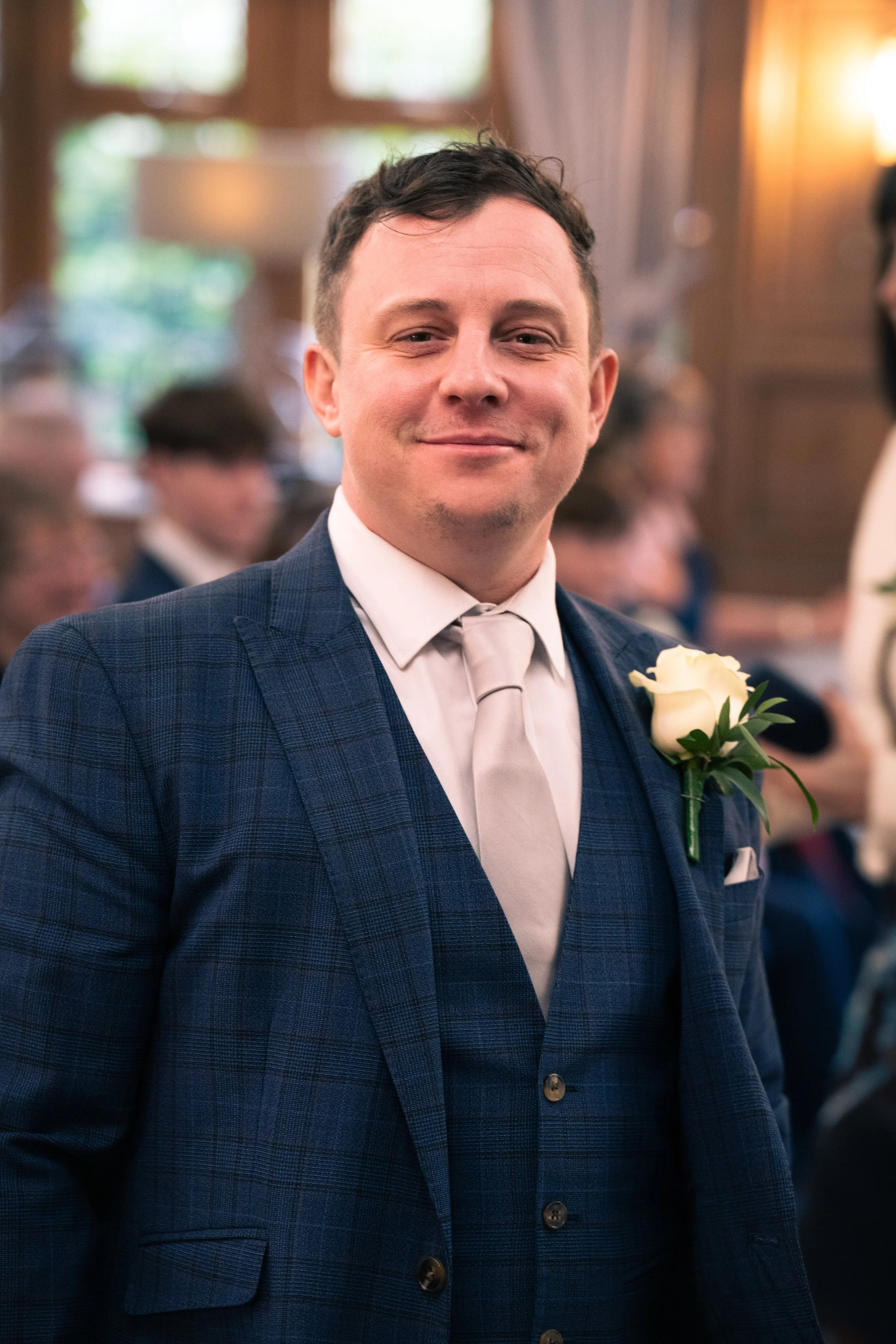 A man in a blue checked suit and white tie, with a white rose boutonniere, smiling at a formal event or wedding reception with blurred guests in the background.