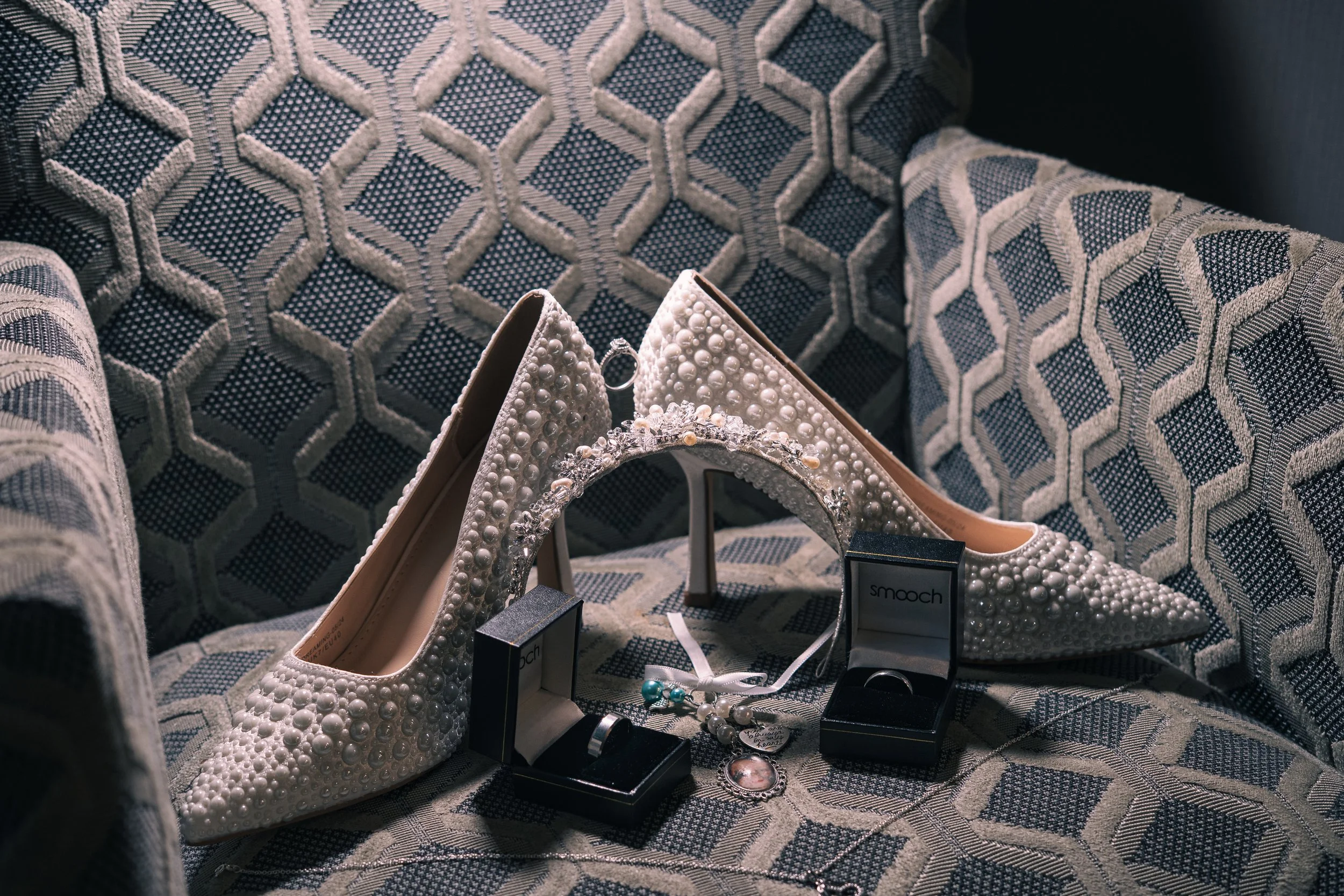 Pearl-embellished white high-heeled shoes with decorative jewelry and rings on a patterned fabric sofa.