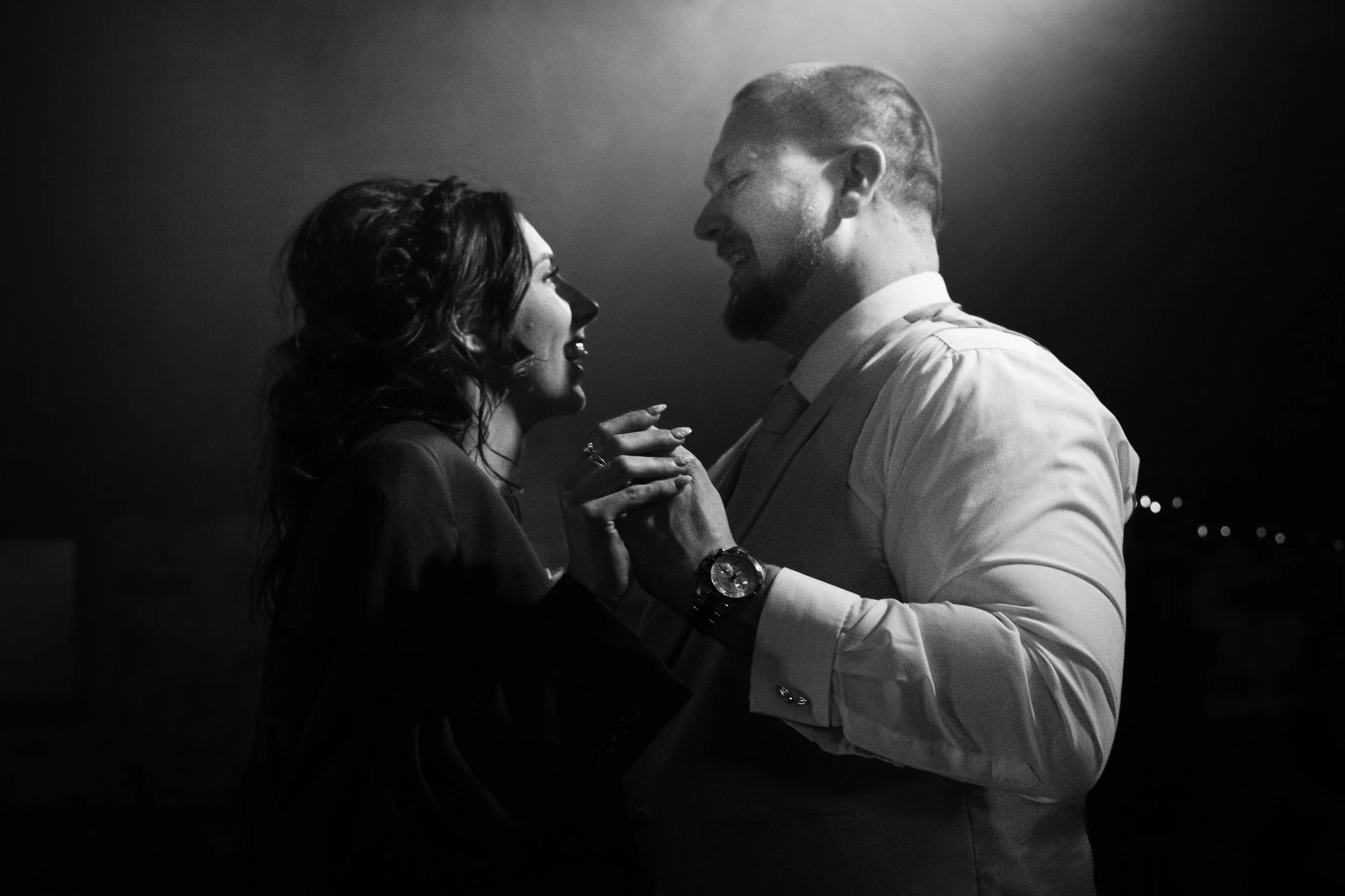 A black and white photo of a man and a woman dancing closely, smiling at each other, with dim lighting and blurred lights in the background.