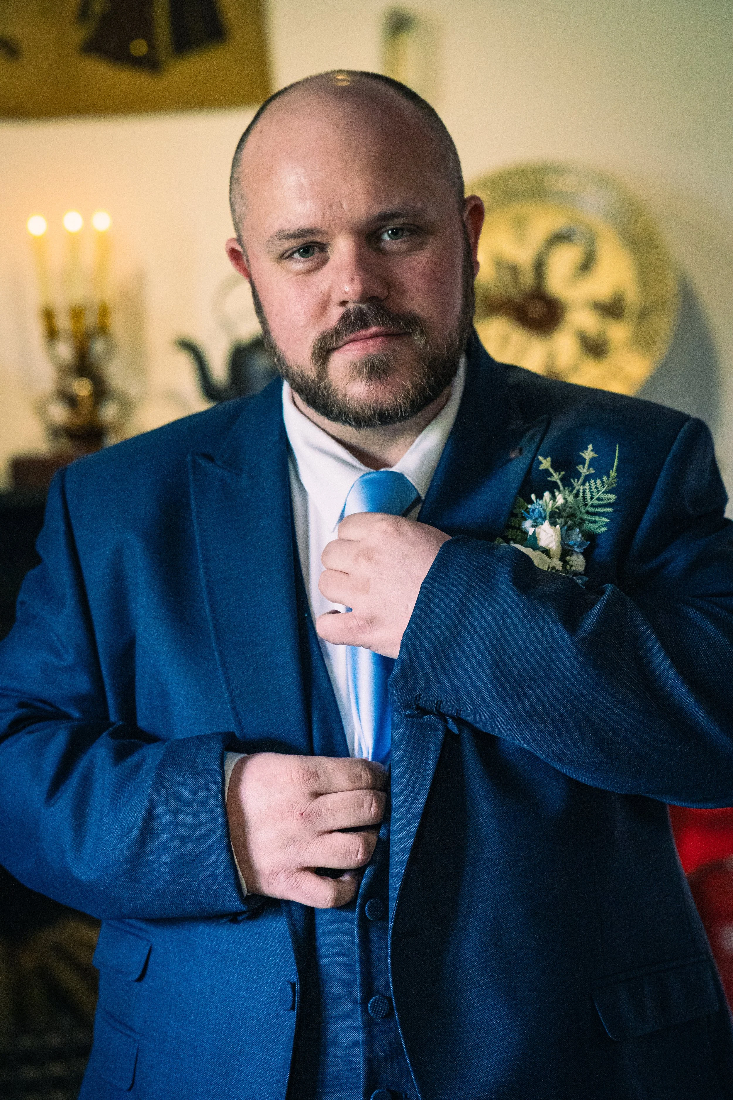 A man in a blue suit adjusting his tie, standing indoors with a blurred background showing a clock and lit candles.