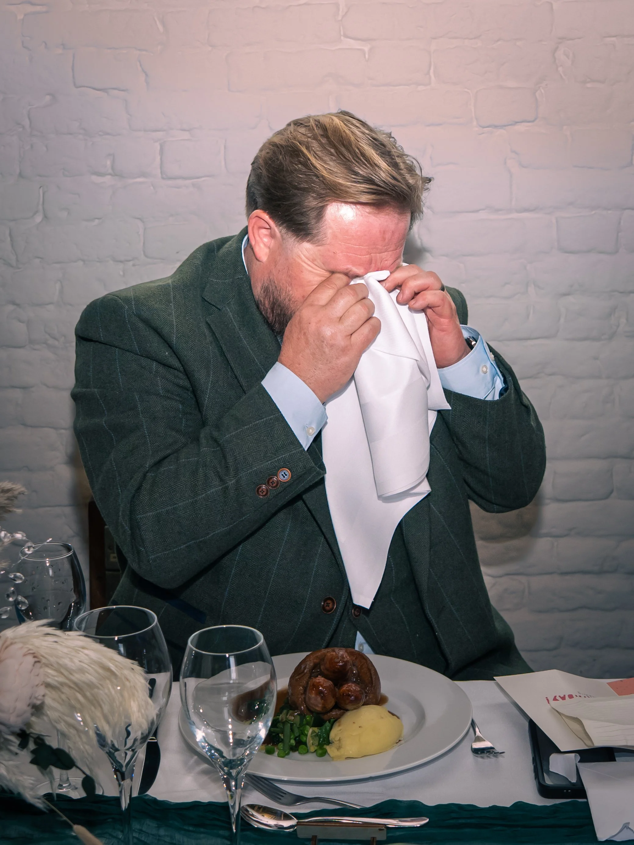 A man in a suit with a beard, sitting at a table, is wiping his eyes with a napkin. The table has a plate of food with mashed potatoes, green vegetables, a large brown cooked meat, and some sausage. There are empty wine glasses, a newspaper, and a wh