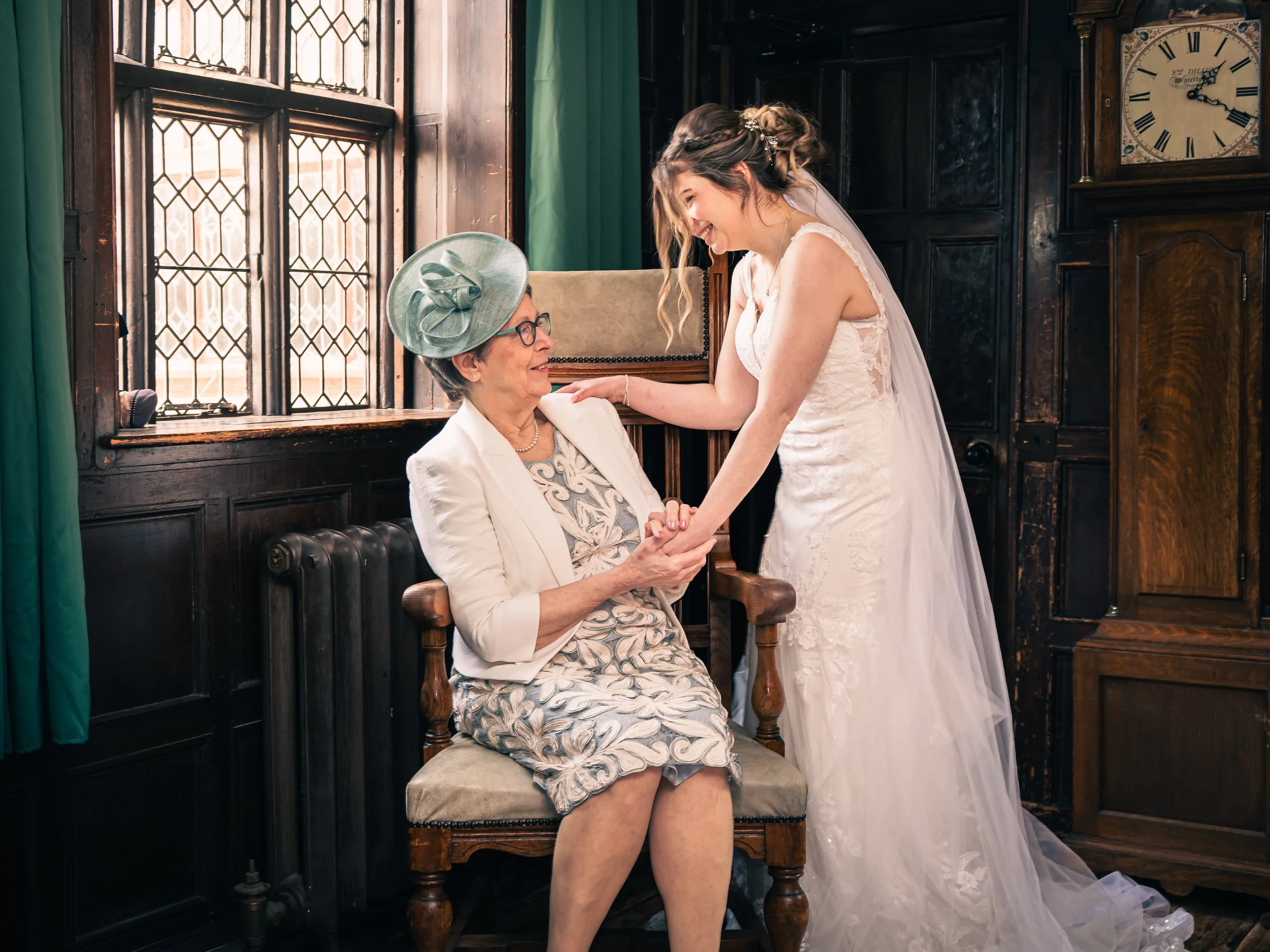 A bride in a white wedding dress and veil smiling and holding hands with an elderly woman in a floral dress, white blazer, and a large green hat, sitting in a wooden chair in a warmly-lit room with dark wood paneling and stained glass windows.
