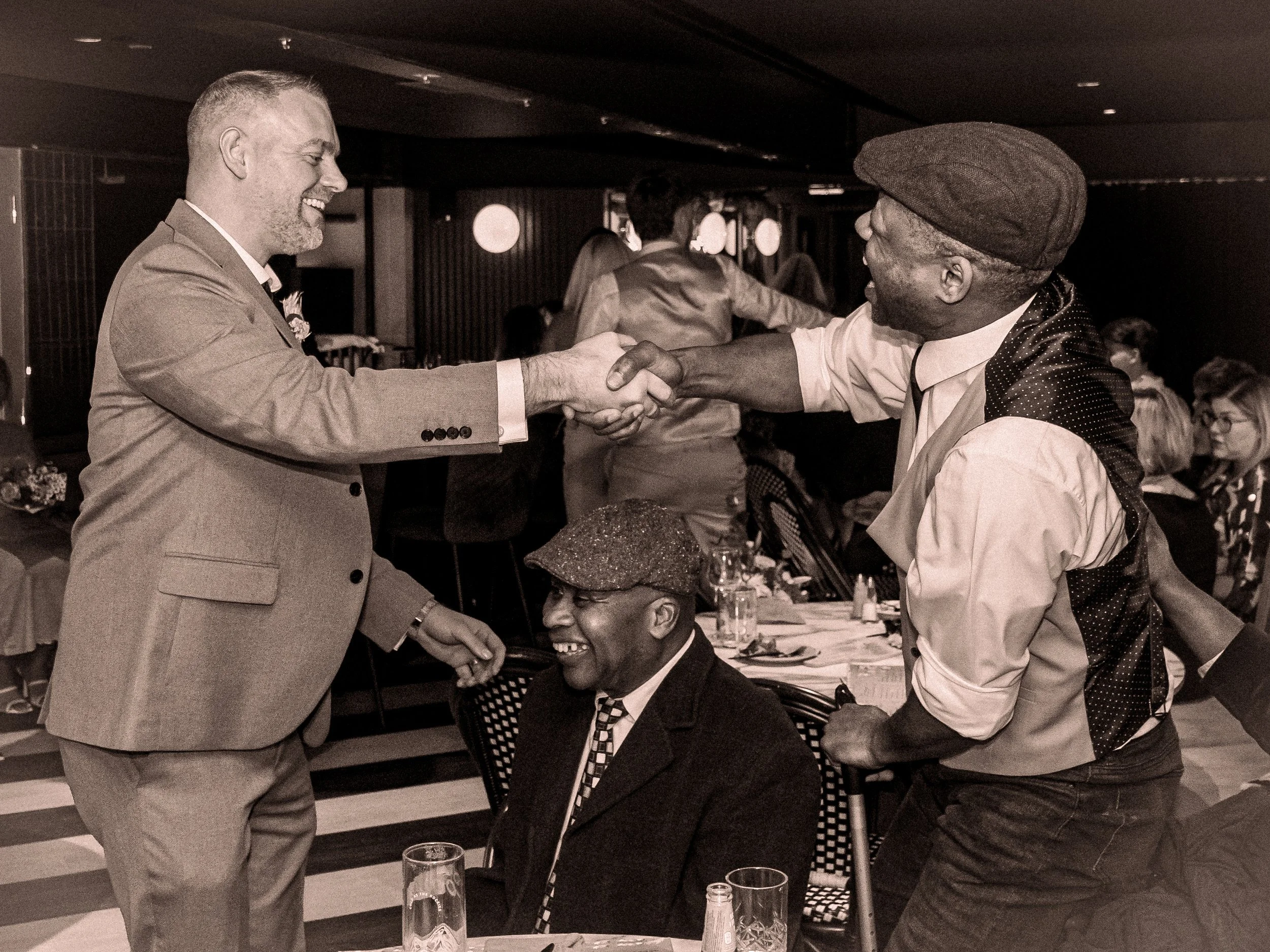 Two men shaking hands and smiling at a social event, with a seated man in the foreground smiling, surrounded by other guests at a dinner gathering.