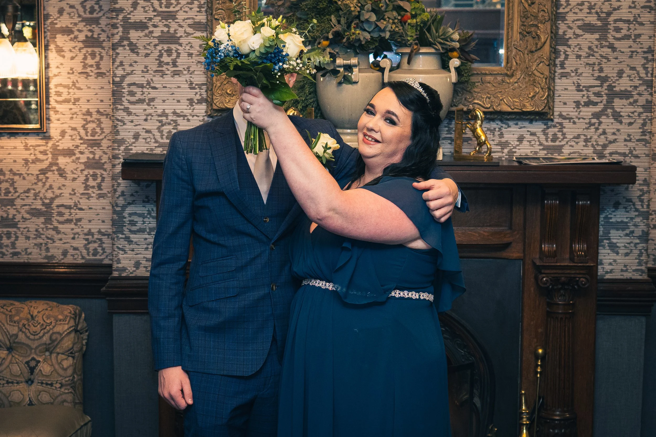 A woman in a dark blue dress and tiara is hugging a person in a blue suit and holding a bouquet of white flowers with blue accents, in an elegant room with a fireplace, floral arrangements, and framed artwork.