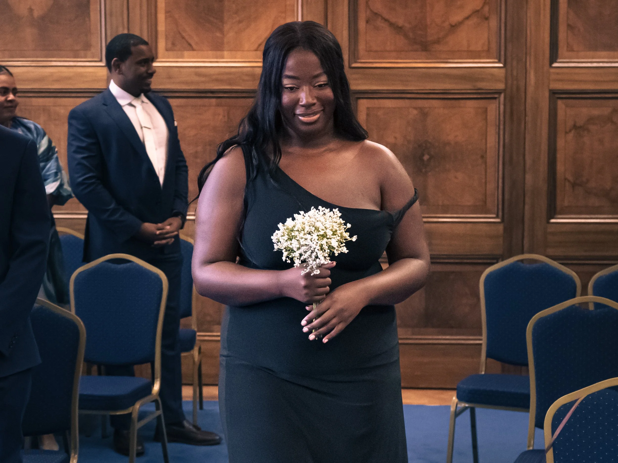 A woman in a black dress holding a small bouquet of white flowers at an indoor event with wood-paneled walls and blue chairs.