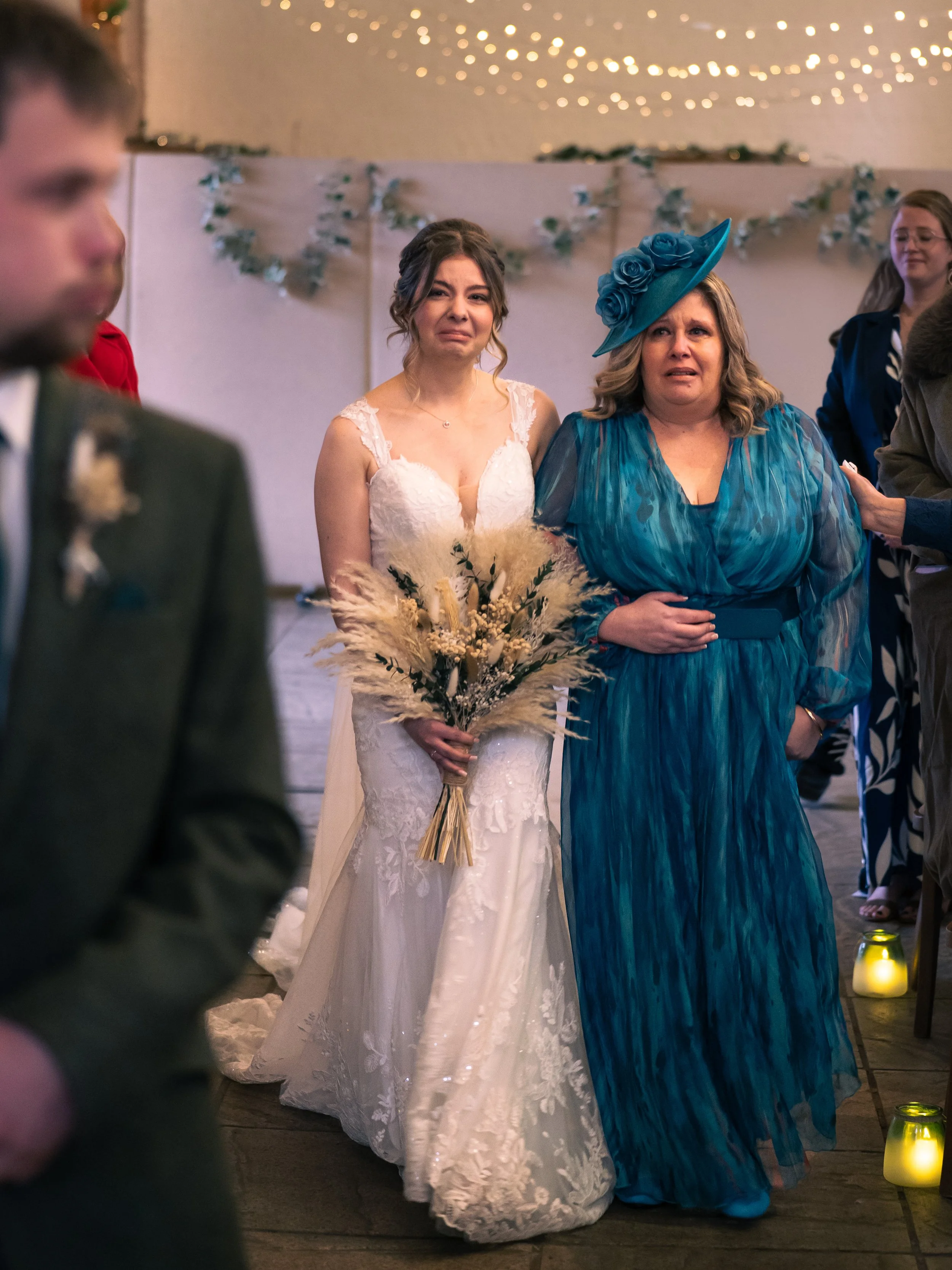 A bride in a white lace wedding dress holds a bouquet of dried flowers while crying during a wedding reception. She is standing next to a woman in a blue dress with a matching hat, also appearing emotional. Other wedding guests are visible in the bac