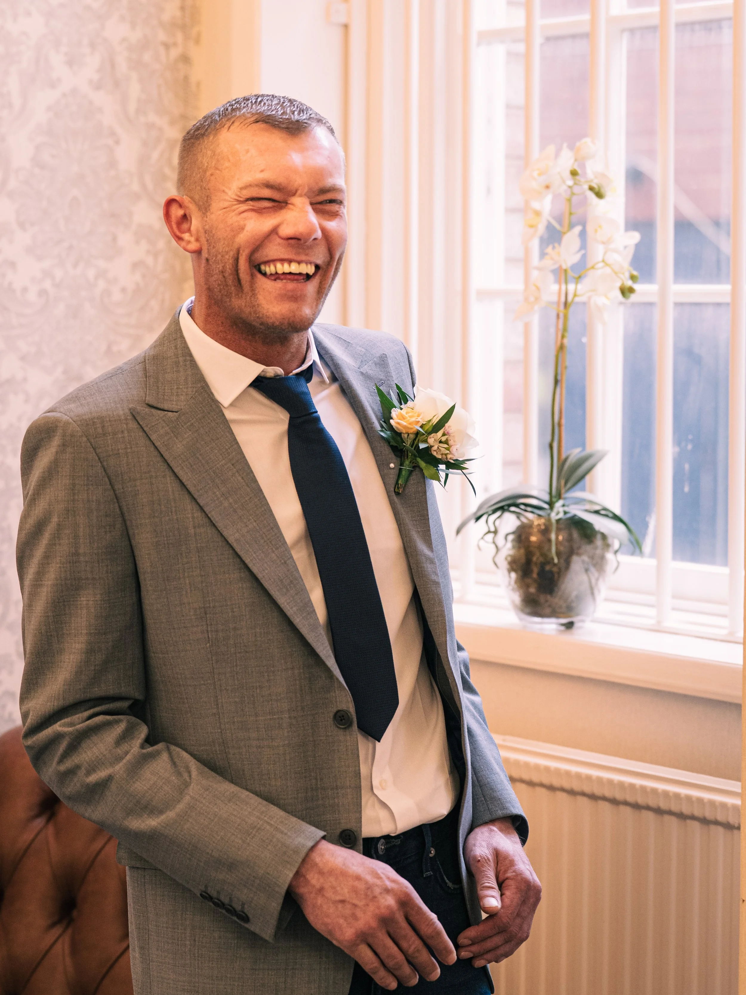 A man in a gray suit and dark tie smiling and laughing indoors near a window with white flowers on the windowsill.