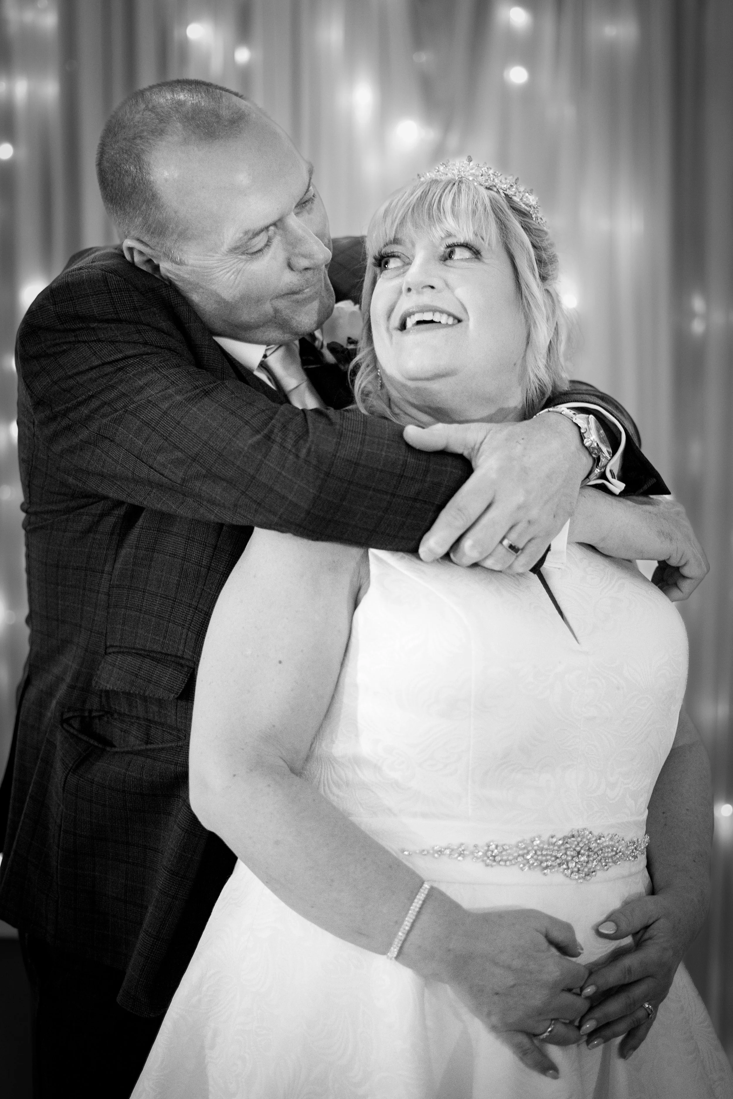 A man in a suit hugging a woman in a wedding dress, both smiling joyfully at each other.