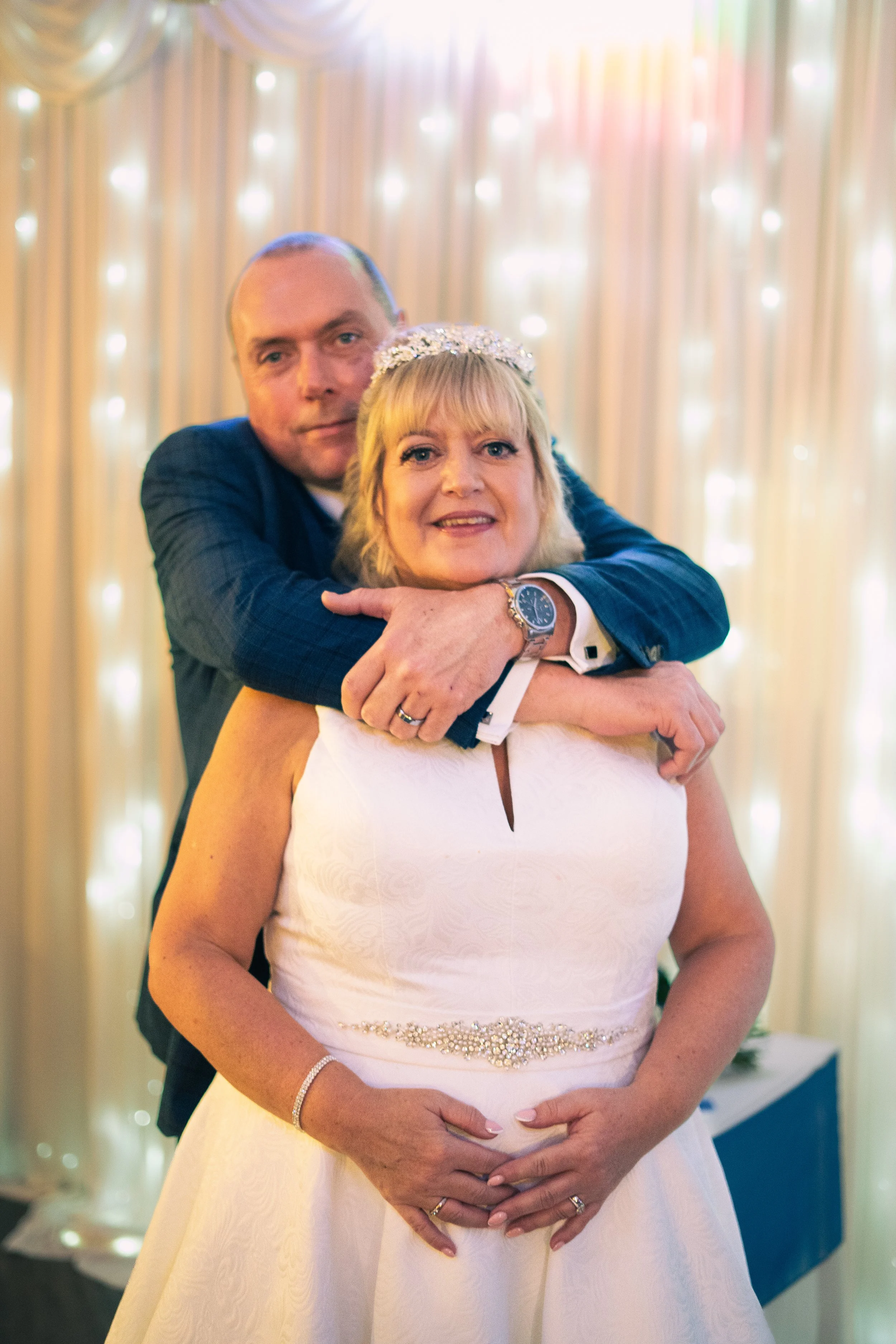 A bride and groom hugging at a wedding reception with lights and curtains in the background.