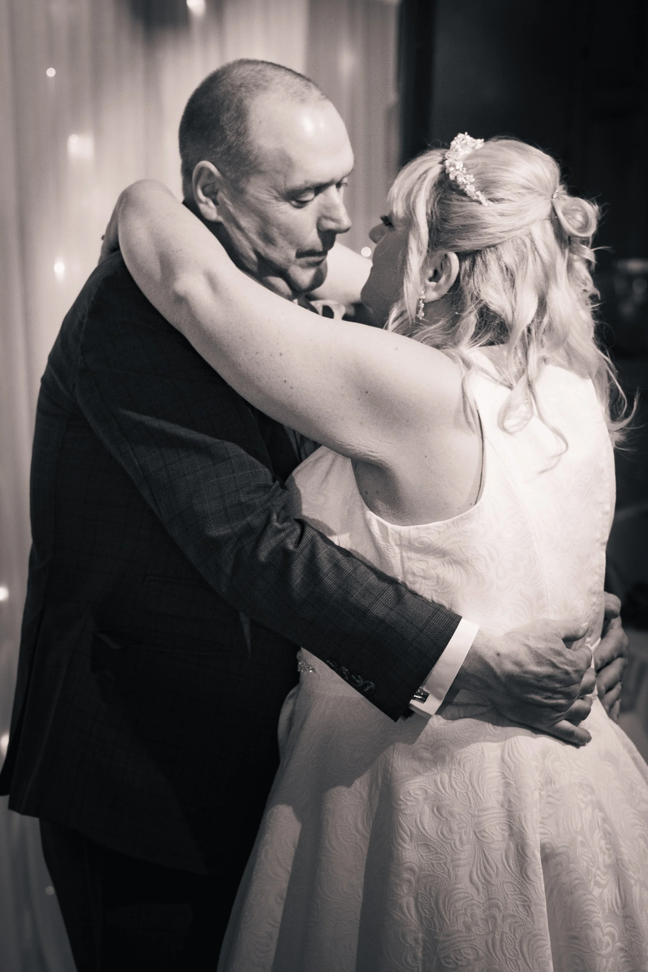 A black and white photo of a bride and groom sharing a dance, embracing closely with her arms around his neck and his hands on her waist, in an indoor setting.