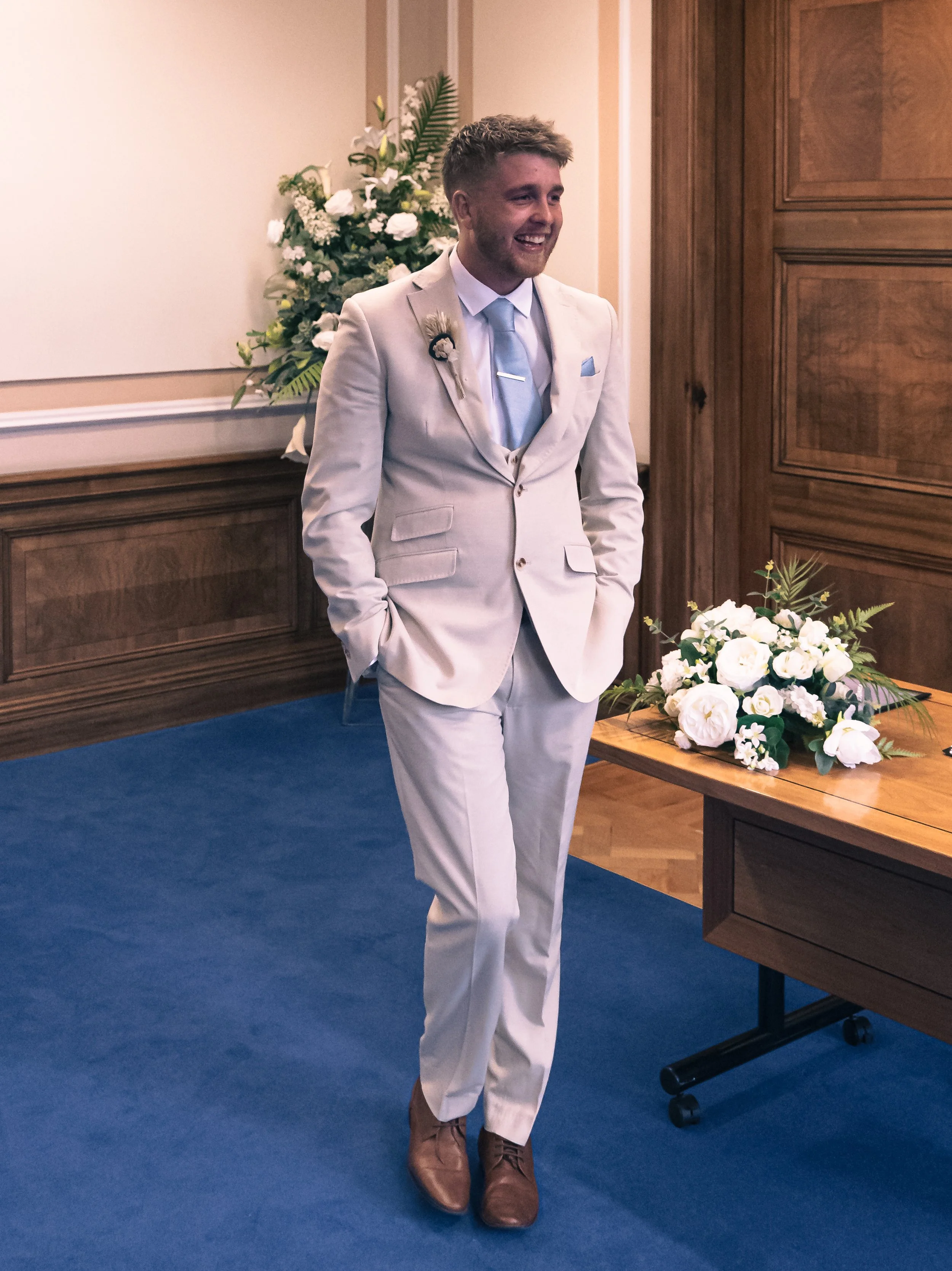 A man in a light-colored suit with a light blue tie and pocket square stands in a room with wooden paneling and floral arrangements, smiling with hands in his pockets.
